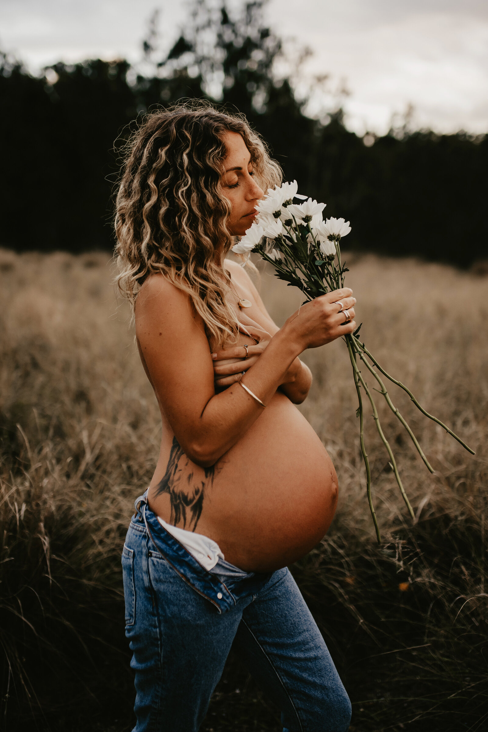 A pregnant person with curly hair holds white flowers and wears blue jeans in an outdoor grassy field, captured beautifully by a family photographer.