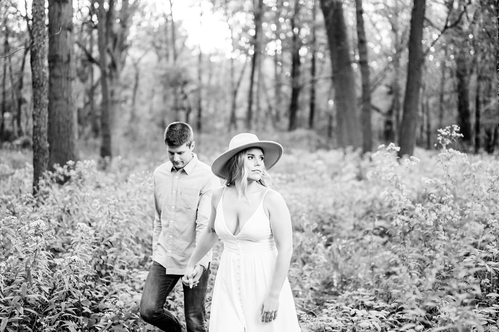 Engaged-couple-walking-through-the-forest-as-the-girl-looks-off-with-her-wide-brimmed-hat