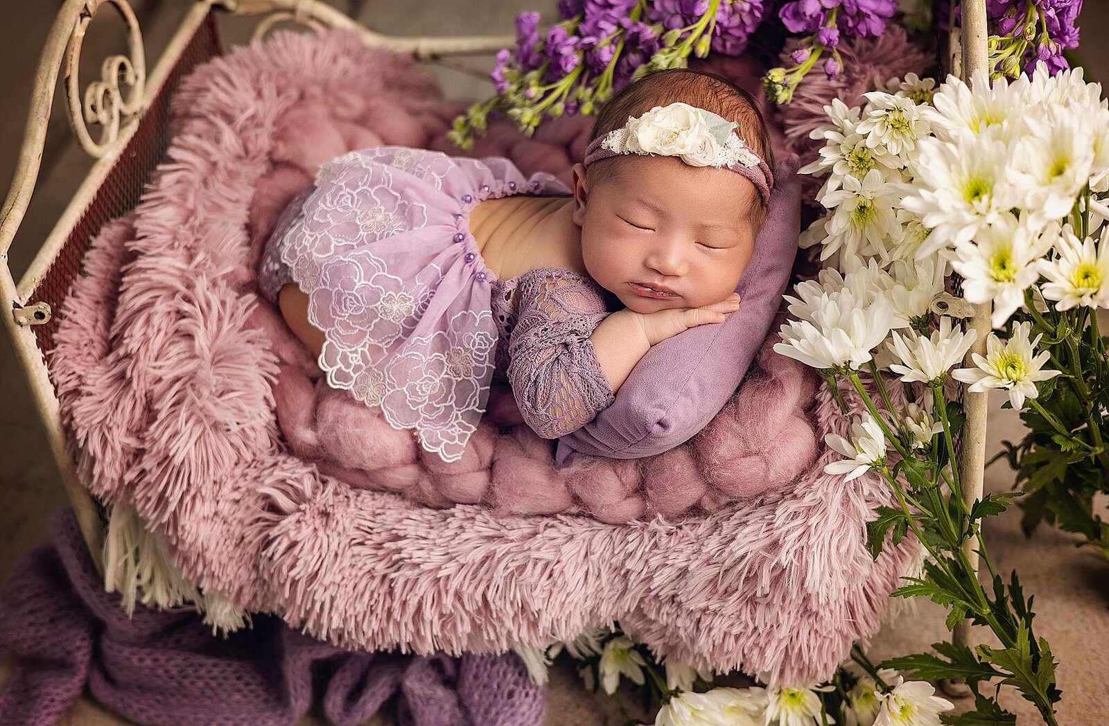 Newborn girl in a purple dress peacefully sleeping on a bed adorned with flowers, captured by Bia Schaefer Photography.
