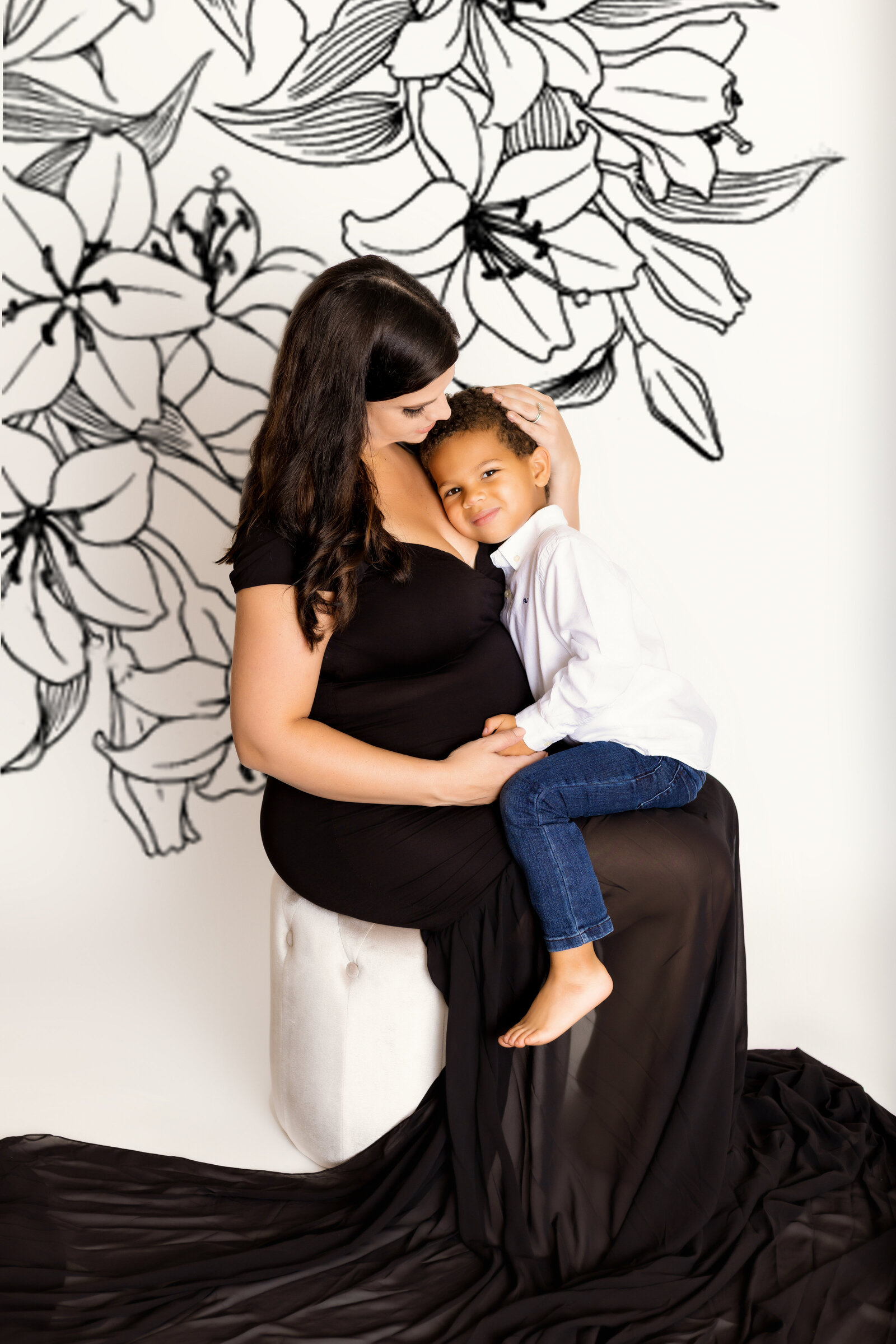 Maternity Photographer, a mother-to-be holds her young son her lap, she wears a black dress