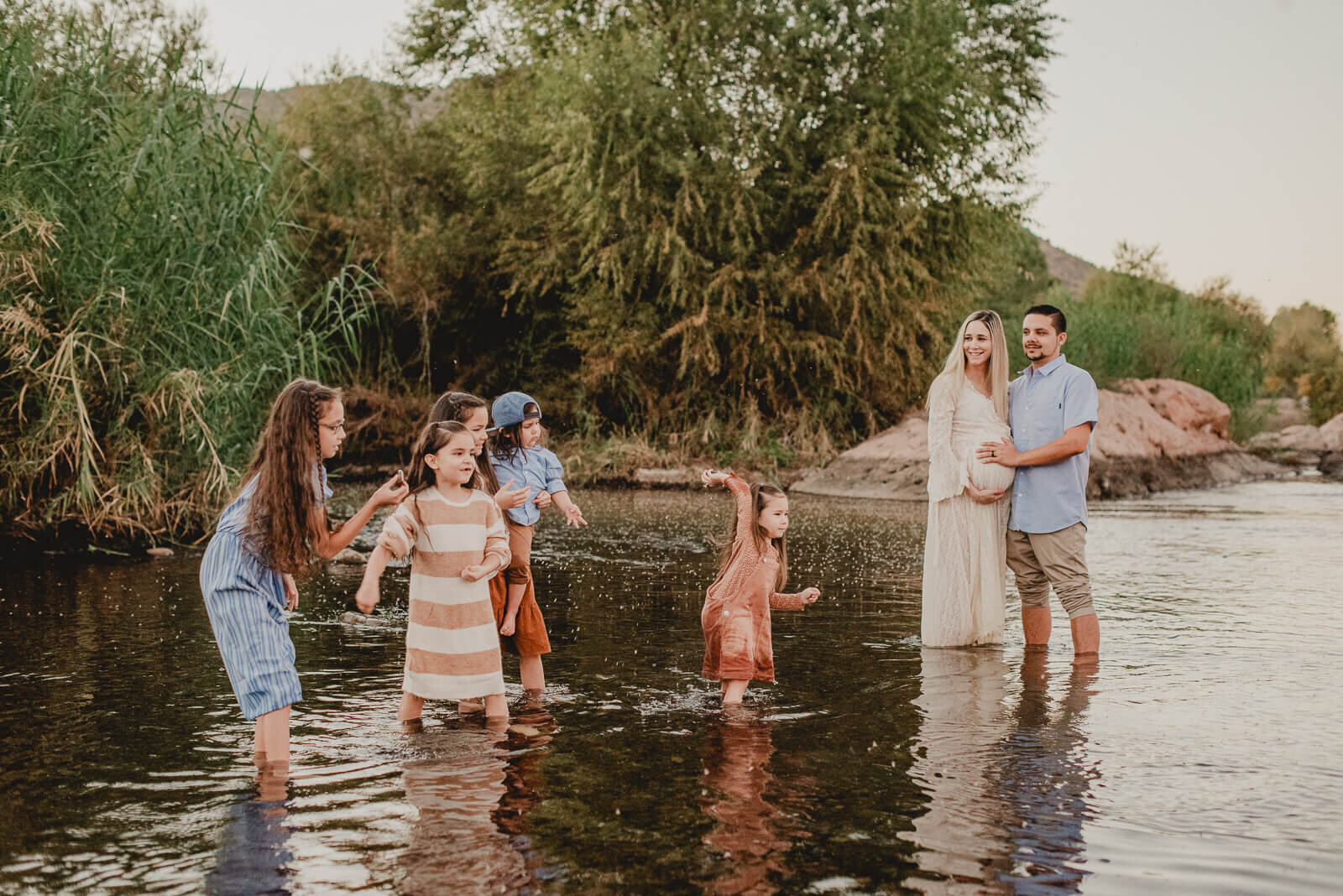 a pregnant mom and her husband look on at their 5 children  playing in the river