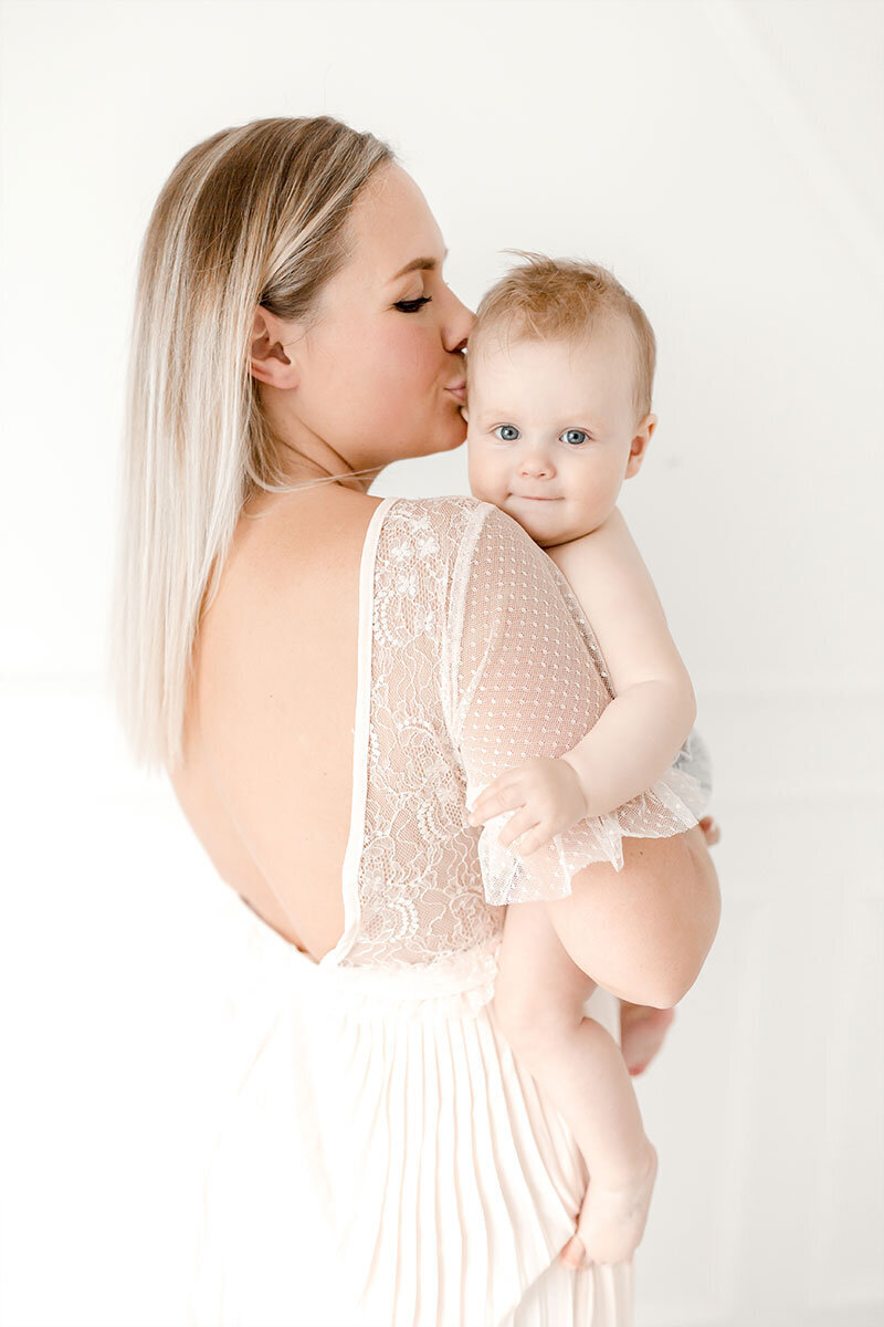 Babyfoto mit Mama. Mama in weißem Kleid hat lächelndes Baby auf dem Arm und gibt ihm einen Kuss auf die Stirn.