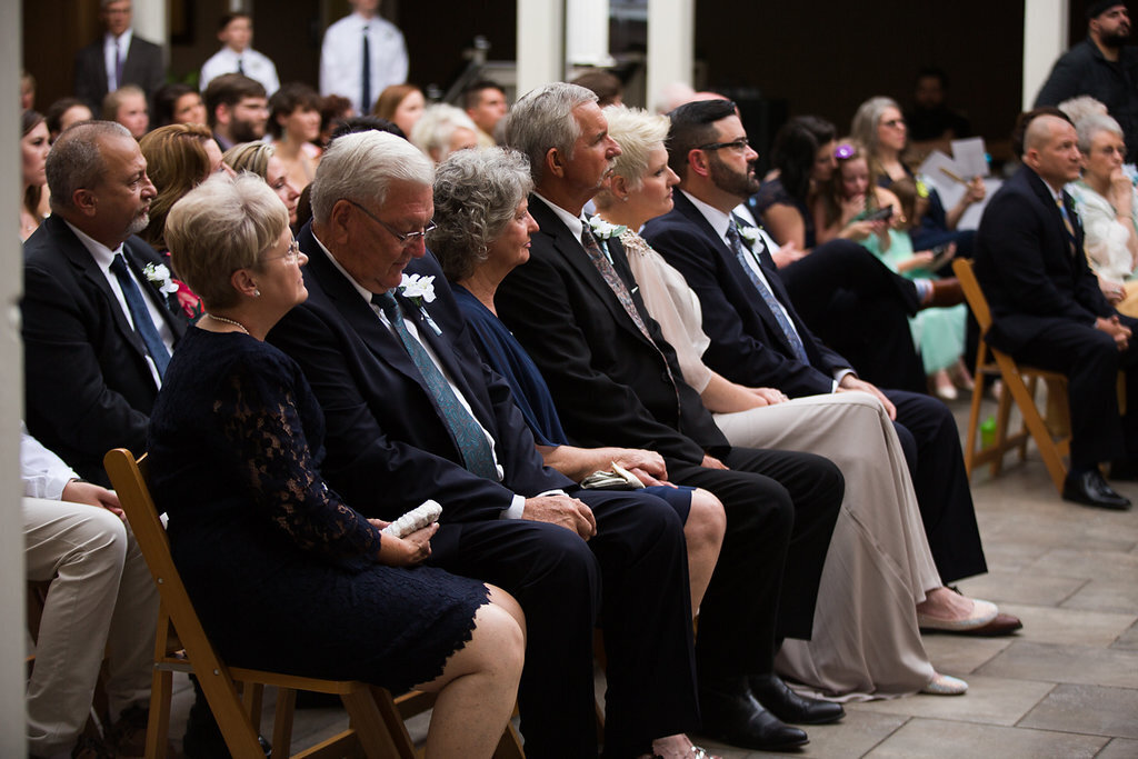 A group of well-dressed guests sit in wooden chairs at an outdoor wedding ceremony, attentively watching the event. More people are seated and standing in the background.