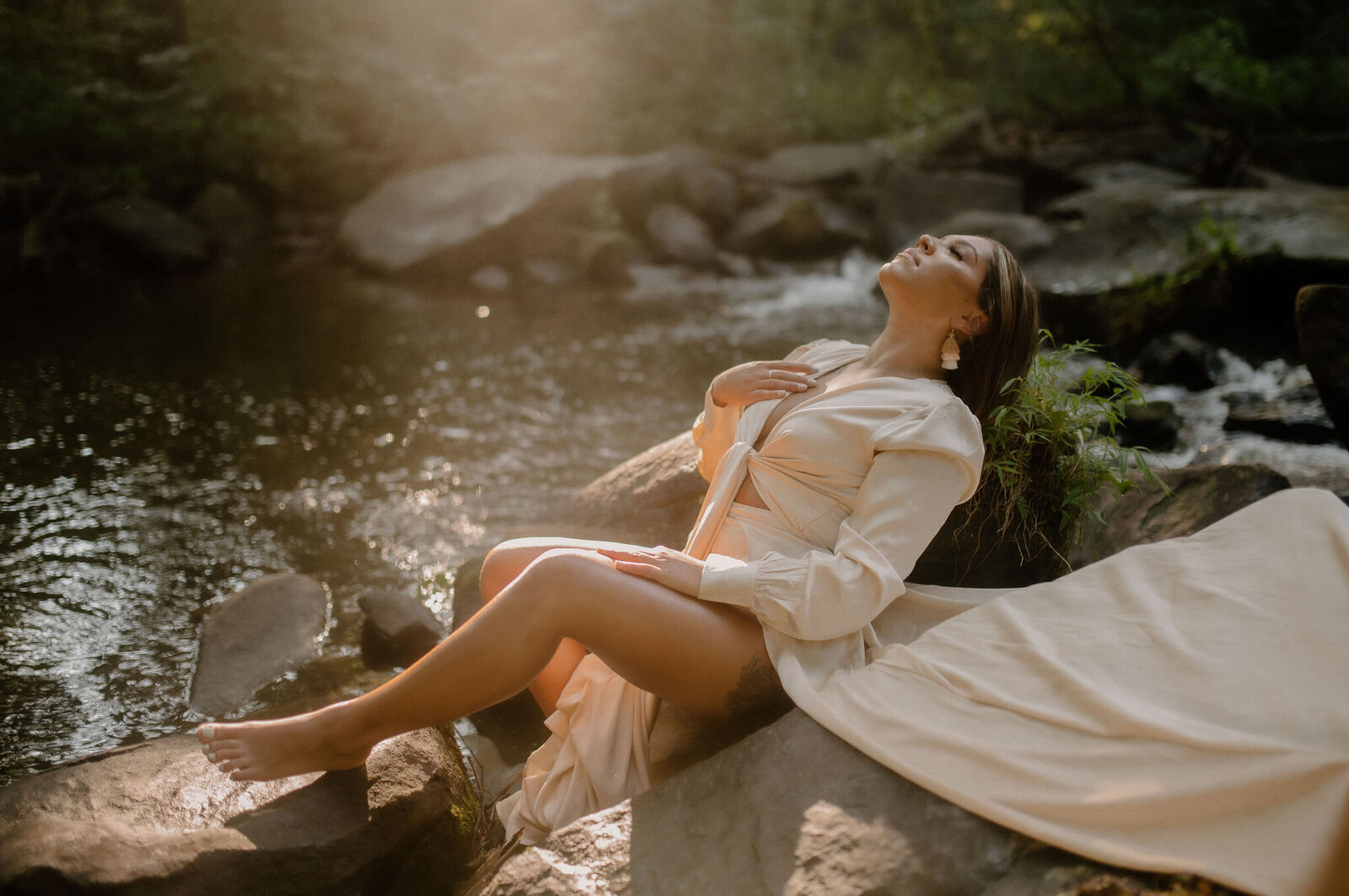 woman in cream dress sitting on rocks in creek peacefully stroking chest with face lifted toward sun