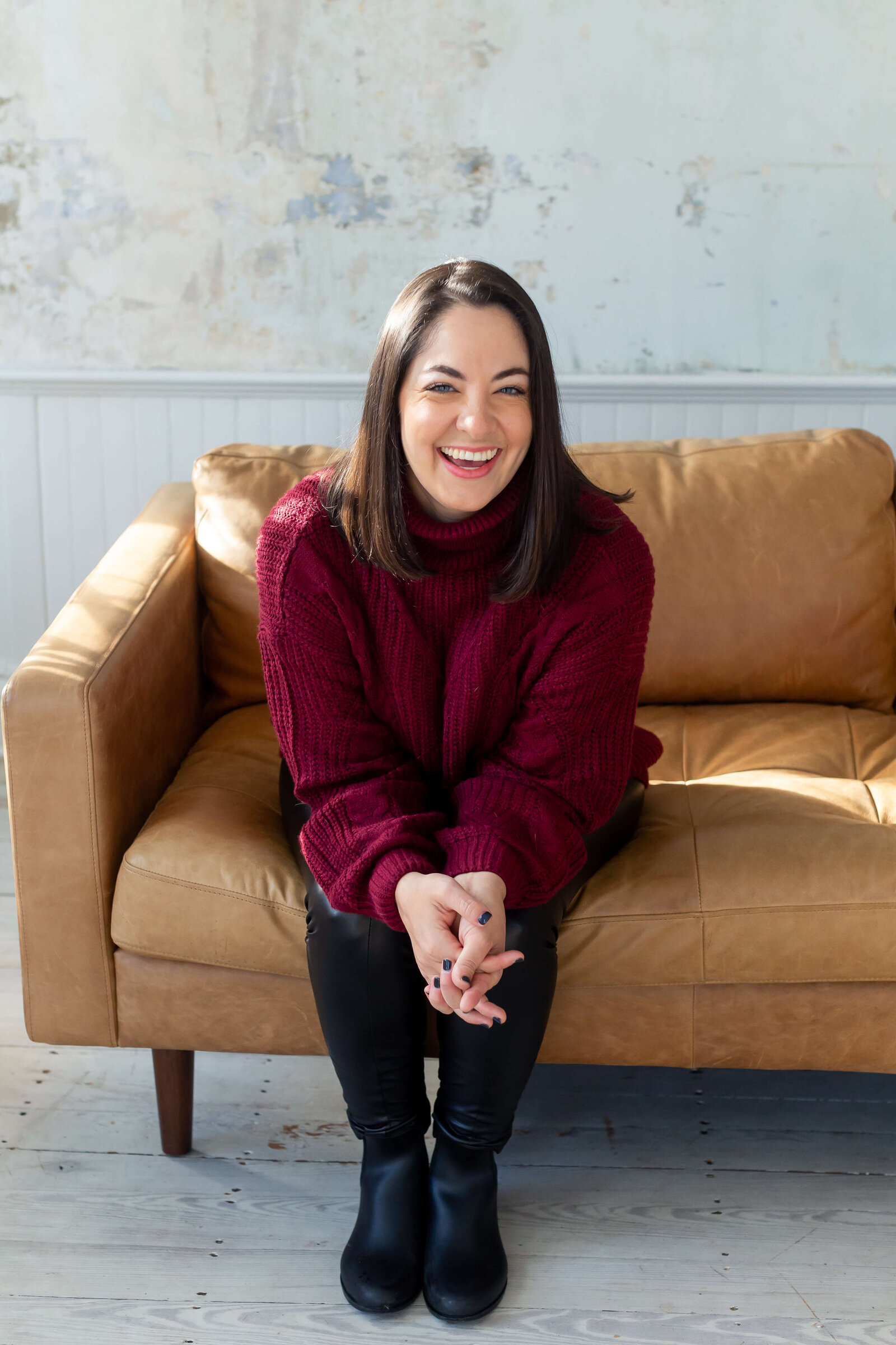 brunette smiling at the camera wearing burgundy sweater sitting on a tan couch
