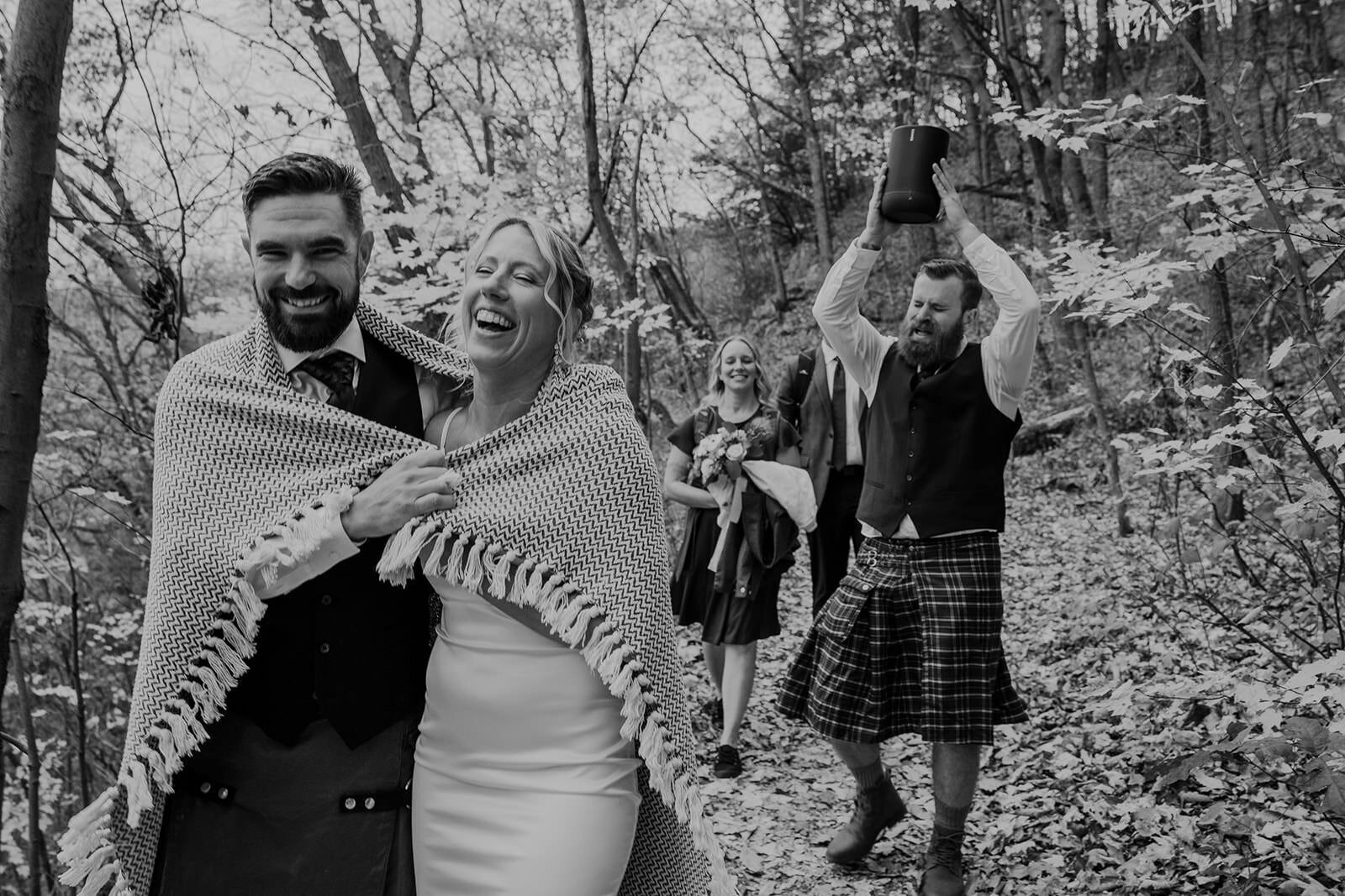 A black and white photo. The bride and groom are walking in front of their friends, and are wrapped together in a blanket. Their friends are walking behind them, and one of them is holding a speaker up in the air, so that everyone can hear the music