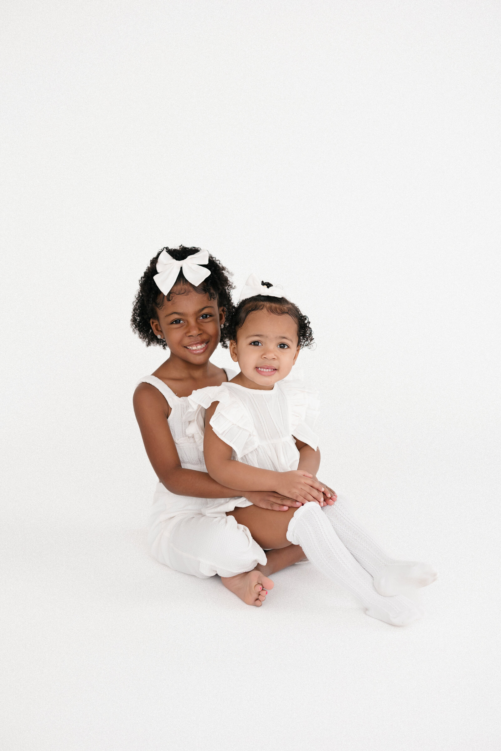 Two sisters wearing all white as they sit on a white backdrop and smile at the camera