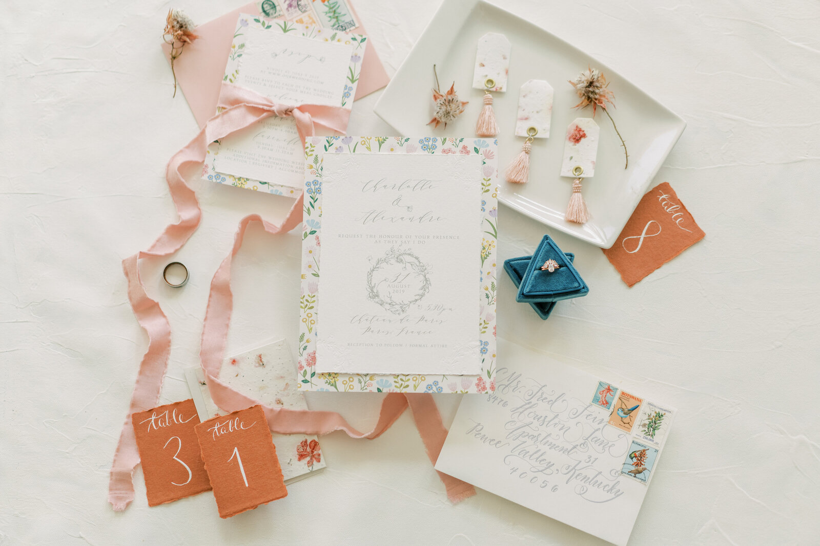Wedding stationery on floral and white card stock with gray font atop a white table with flowers, a jewelry box, and ribbon.