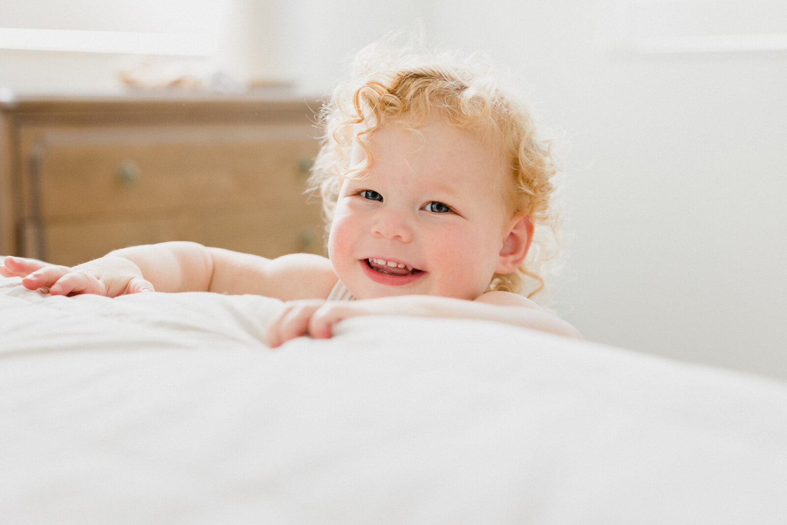 A curly haired toddler girl smiles at the photographer while trying to climb on her mom and dad's bed