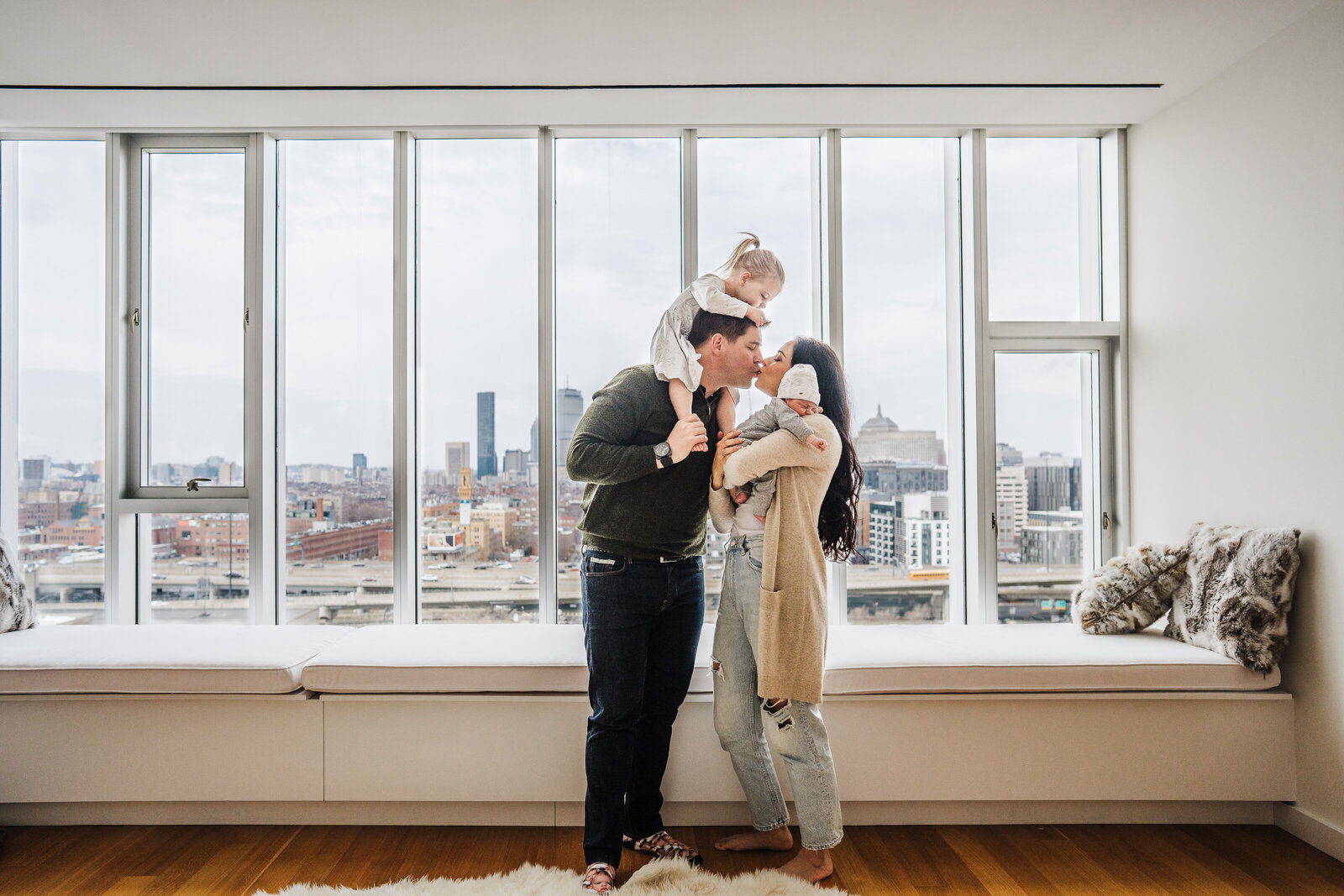 parents hold toddler and newborn in bedroom overlooking the boston skyline