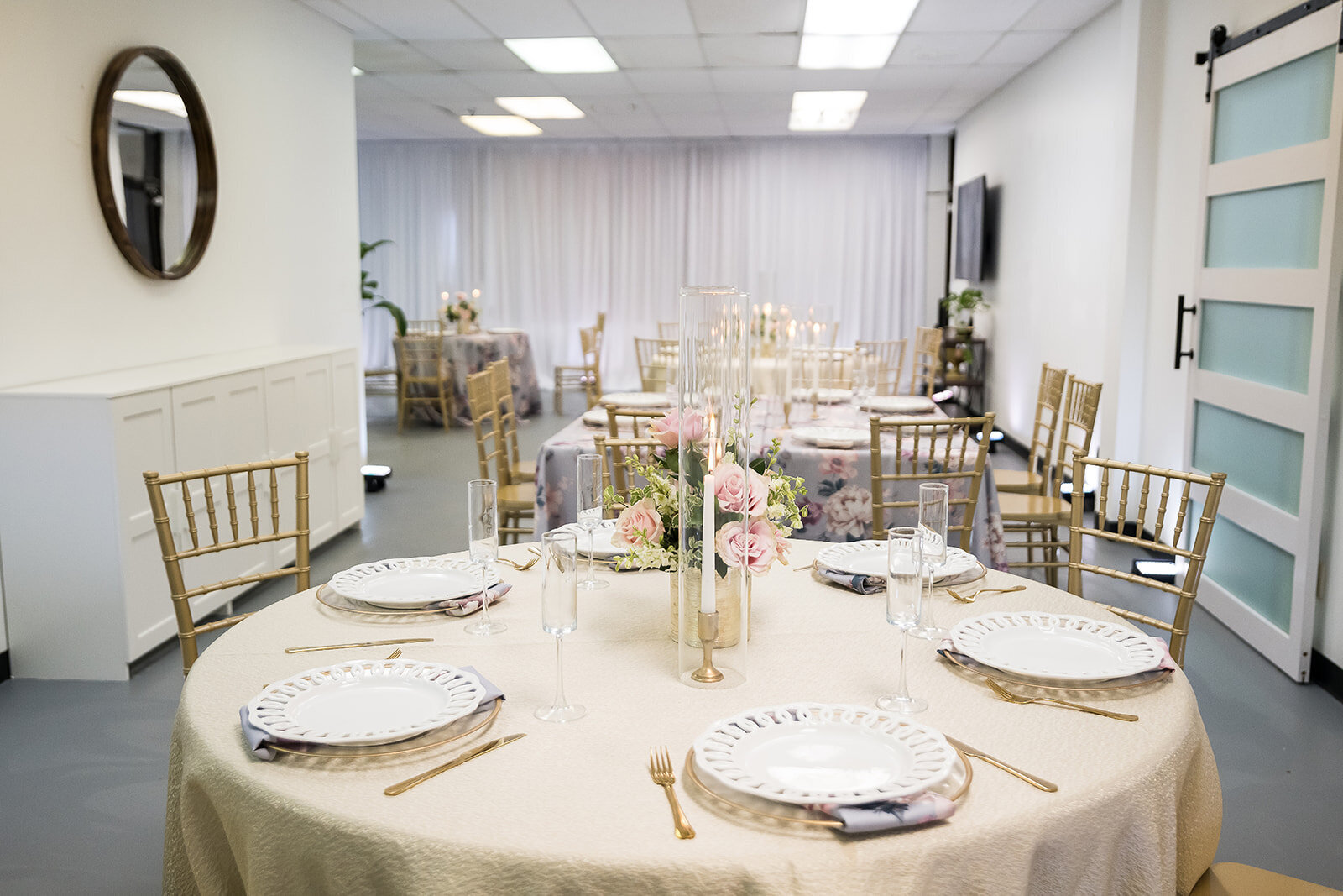 View of The Parlor at Wells set for a seated event with soft blush and floral accents, featuring romantic candlelight, gold chairs, and white draping in a bright and airy venue.