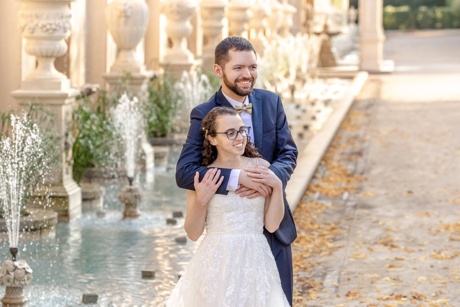 Romantic portrait of a bride and groom embracing by a fountain in a historic Longwood Gardens in Pennsylvania, surrounded by elegant architectural details and vibrant summer foliage.
