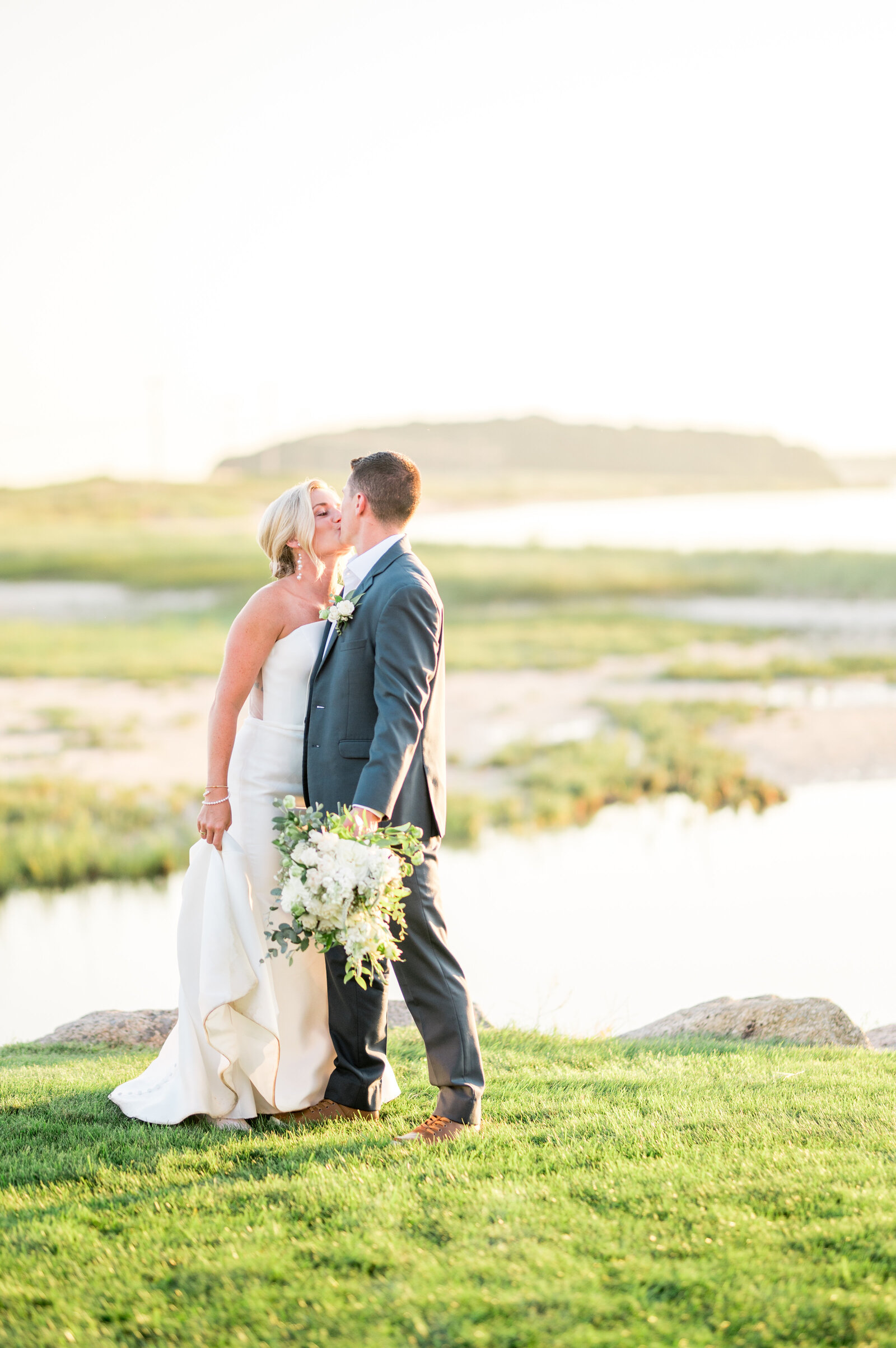 Bride and groom sharing a kiss on a coastal lawn in Bourne, MA during golden hour, holding a soft white bouquet with the shoreline glowing behind them — warm, dreamy, and coastal romance at its finest.