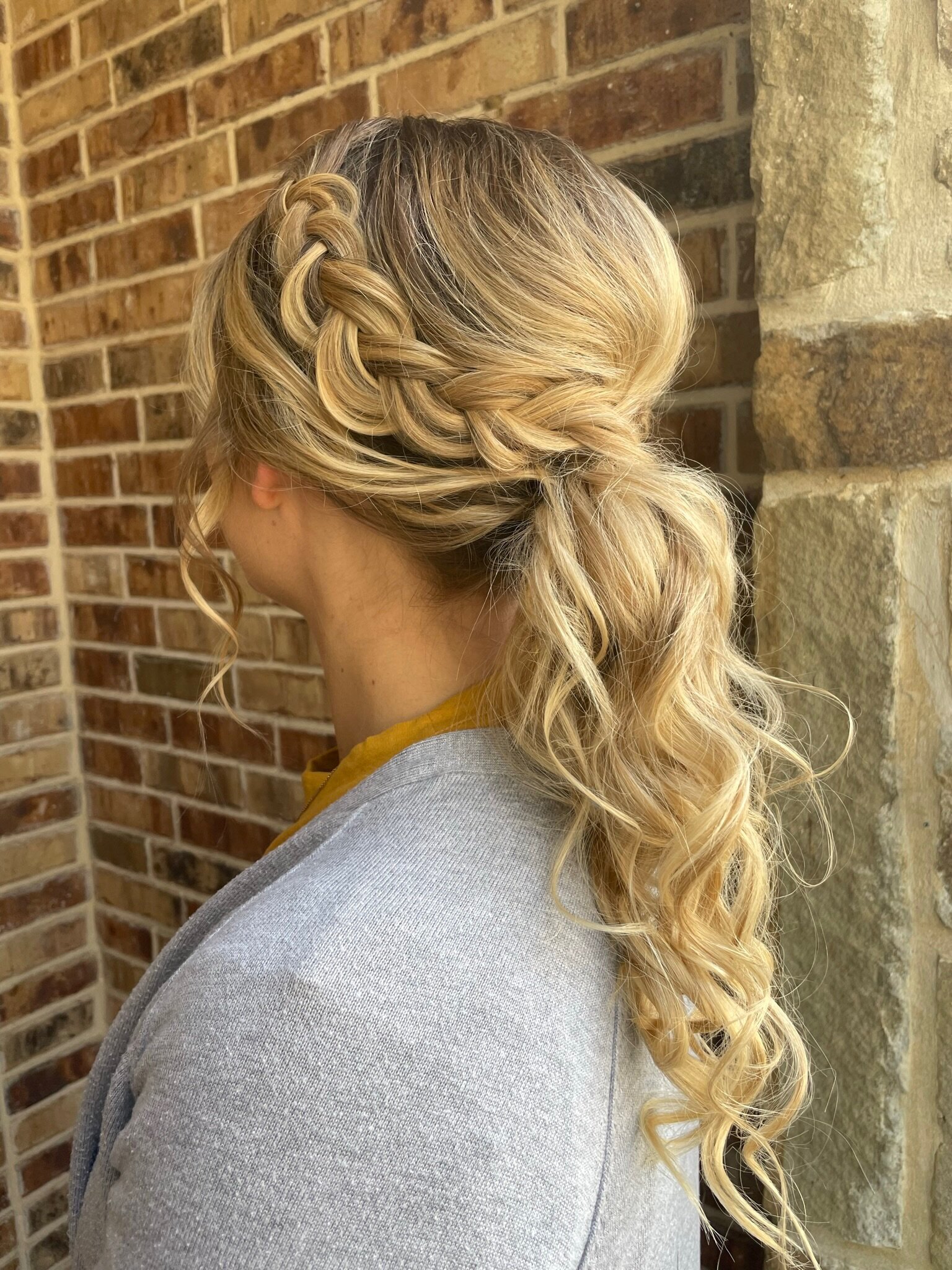 A woman with braided blonde hair styled in a wavy ponytail, wearing a gray sweater, poses against a brick wall. Her look perfects what to expect from a wedding makeup artist, blending elegance into any bridal beauty experience Texas has to offer.