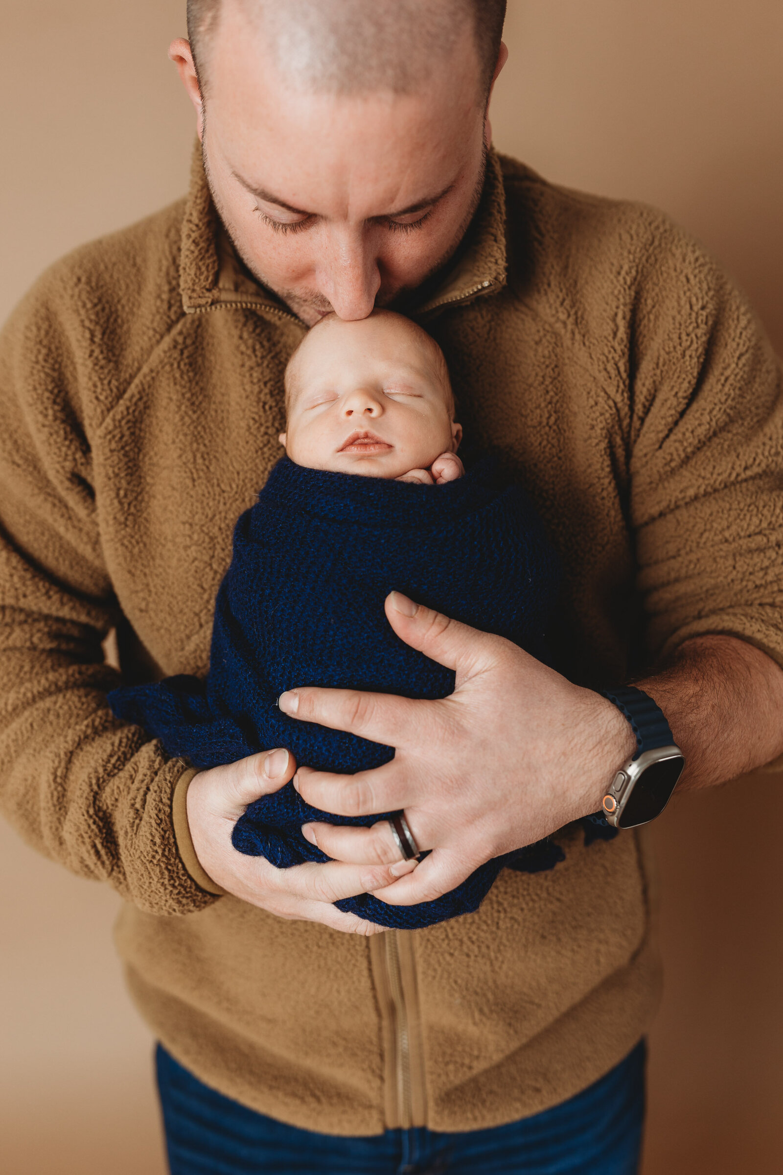 Dad in brown fuzzy sweater holding swaddled newborn baby kissing the top of his head