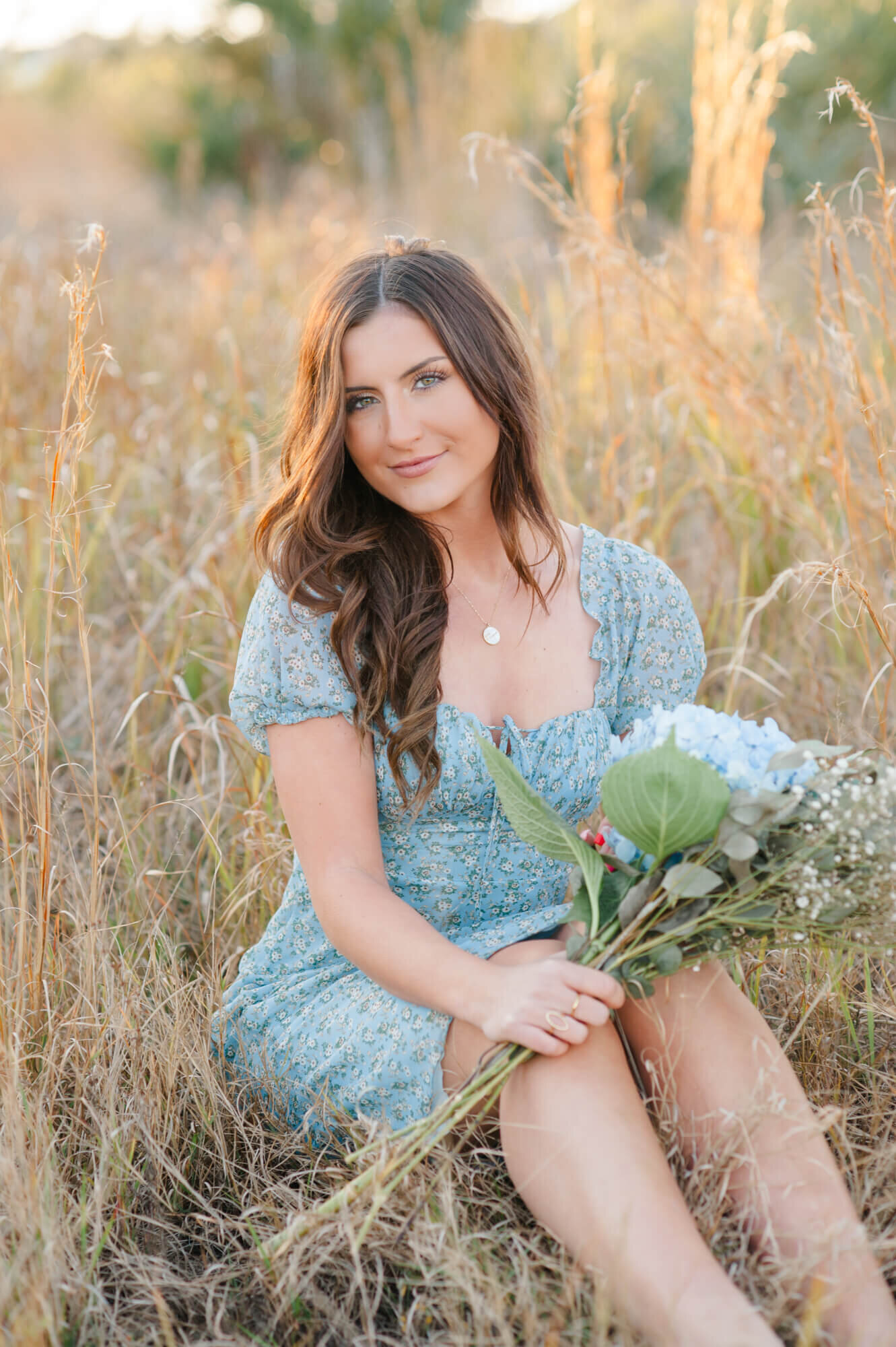 Senior girl sitting in tall grass at sunset sitting in a blue dress holding florals