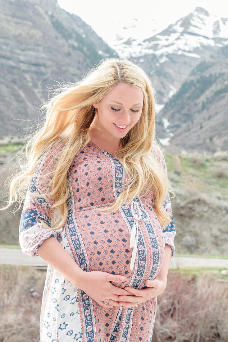 Utah maternity photography of a pregnant woman looking down at her belly with mountains behind her. Photo taken at Provo Canyon in the spring