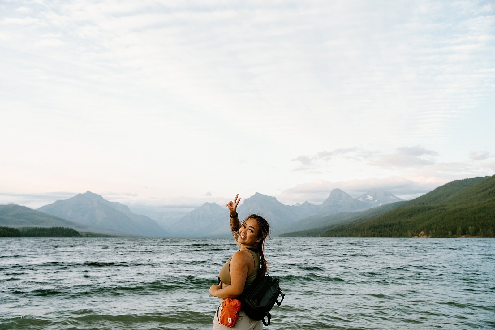 woman twisting upper torso to look back to the camera while throwing a peace sign up with her right hand with water and mountains as her background