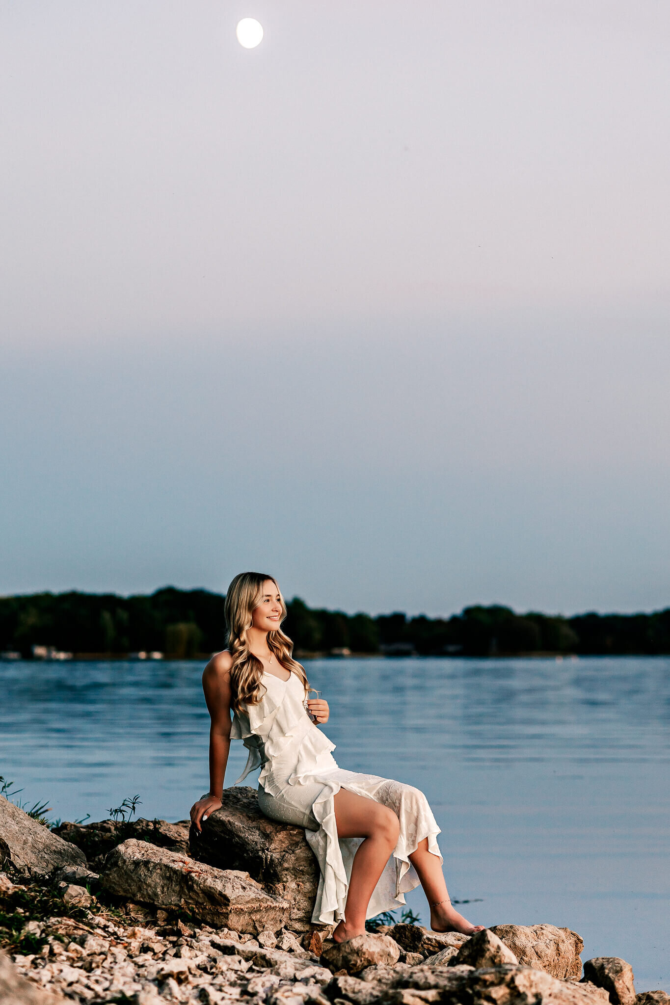 A woman in a white dress sits on rocks by a calm lake under a clear sky with a visible moon. The background shows distant trees and the peaceful water reflects the evening light.