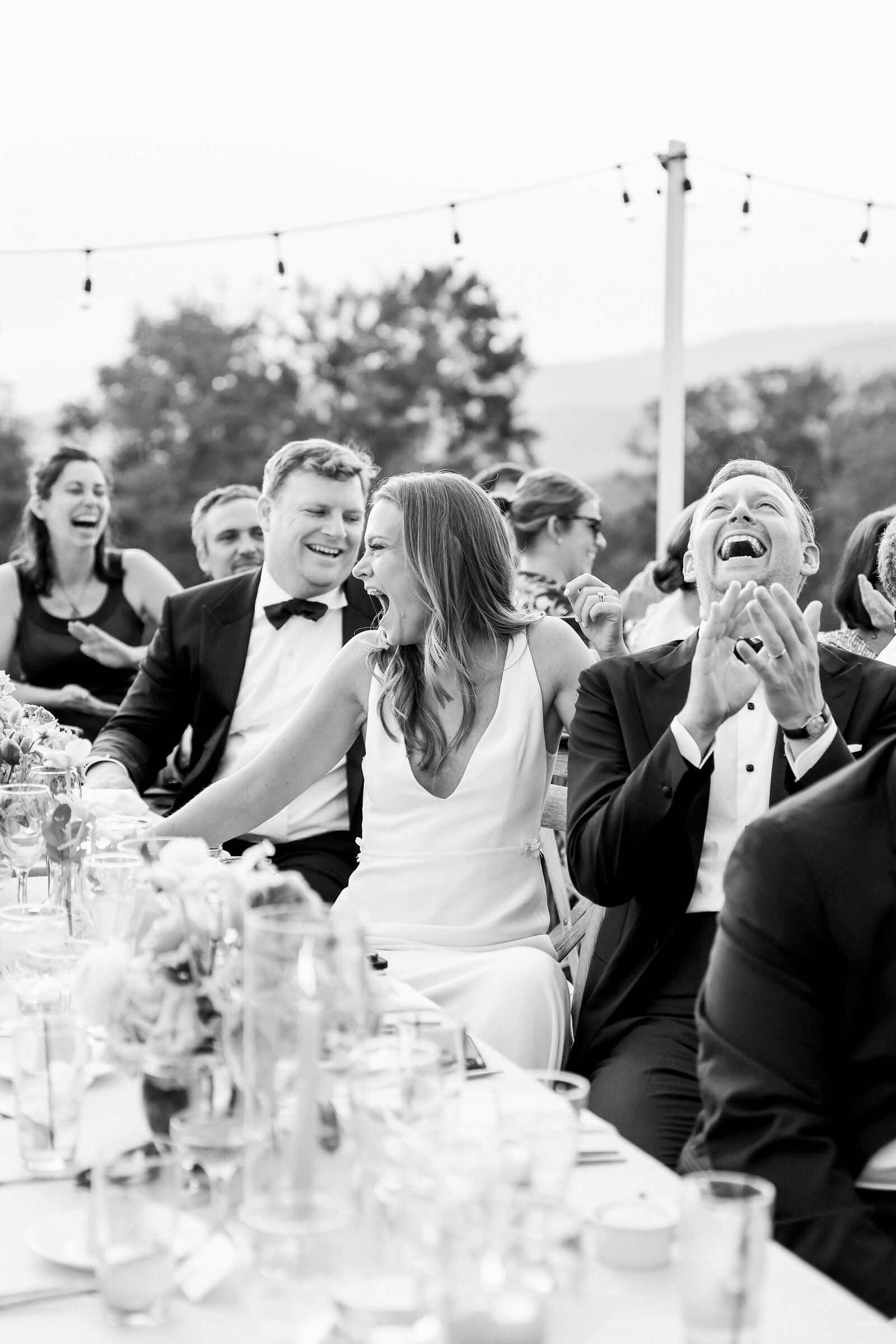 Wedding couple and guests laughing and clapping as they sit at a table at a reception.