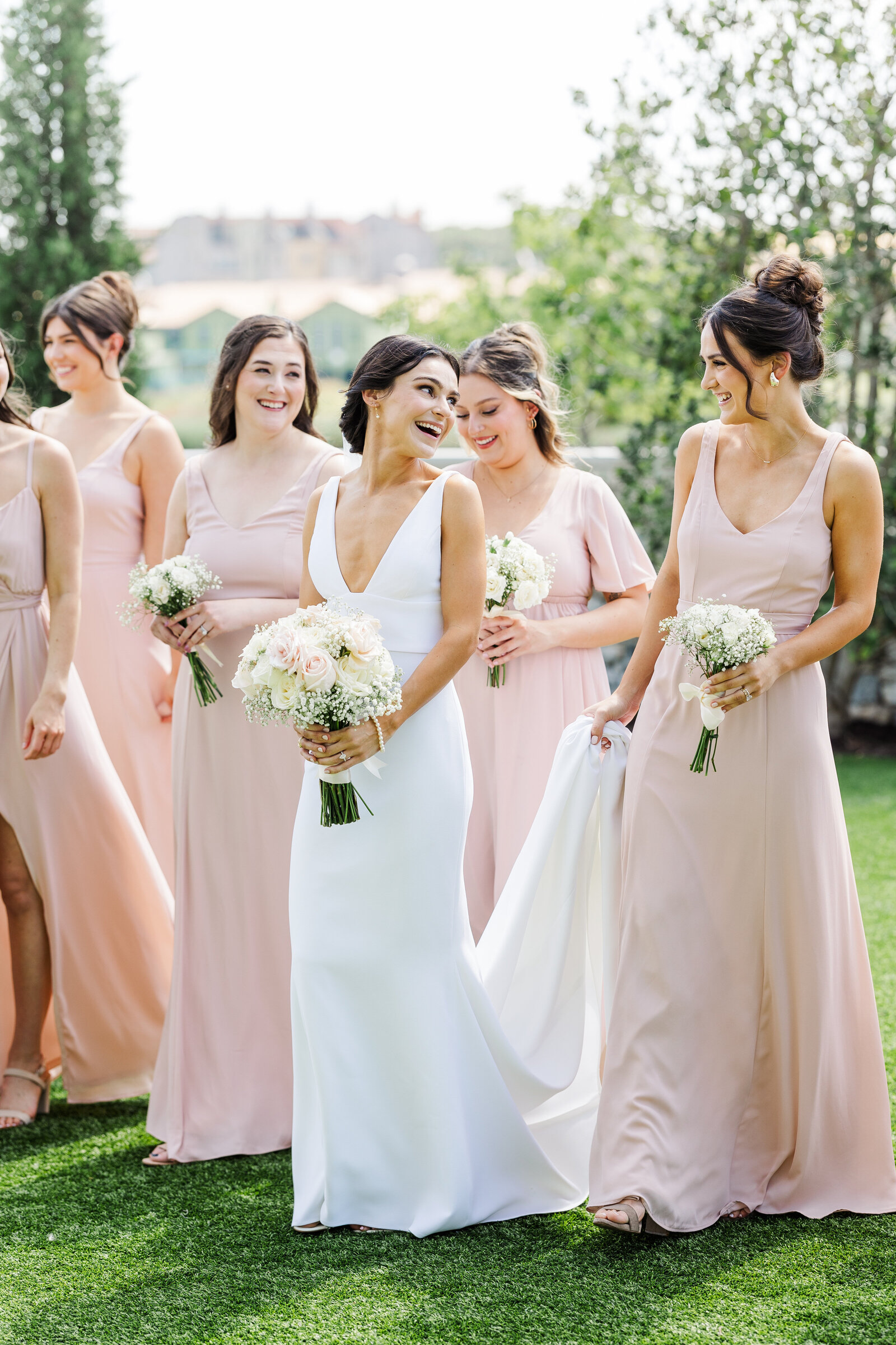 Happy bride in a white gown laughing with her bridesmaids dressed in blush pink dresses, holding white rose and baby’s breath bouquets, captured in an outdoor garden setting with soft natural light.