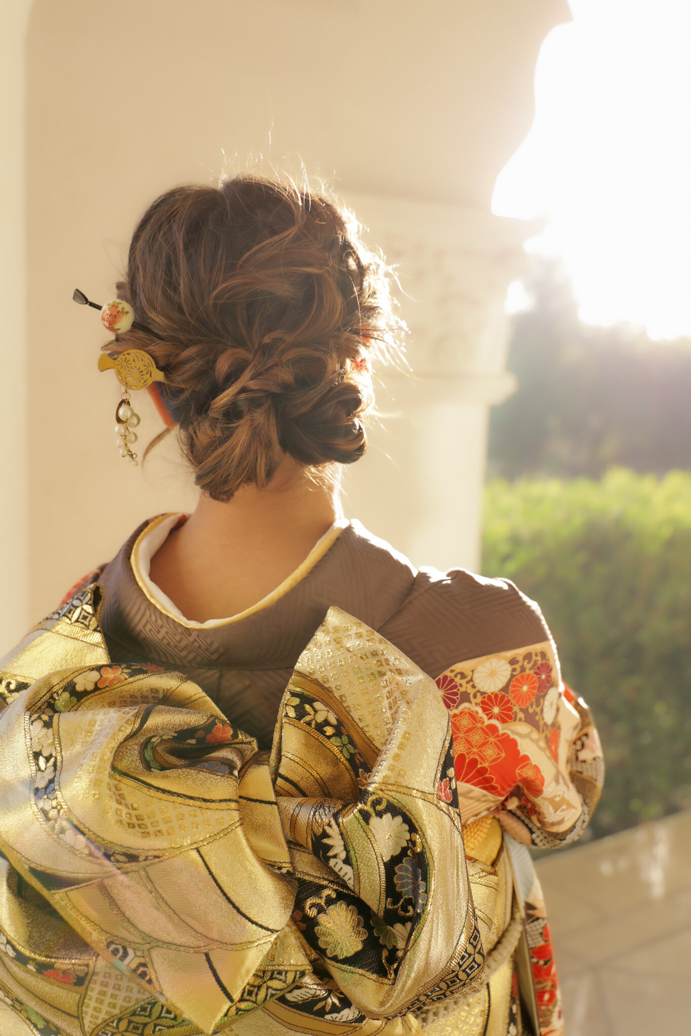 Back view of an intricate updo with decorative kanzashi hair ornaments, highlighting the obi bow of a brown furisode at Brand Park Los Angeles during a Seijinshiki celebration. Girl’s kimono rental in Los Angeles | ブランドパークで成人式を祝う振袖姿、華やかな帯結びと髪飾りを後ろから。ロサンゼルスで女子着物レンタル
