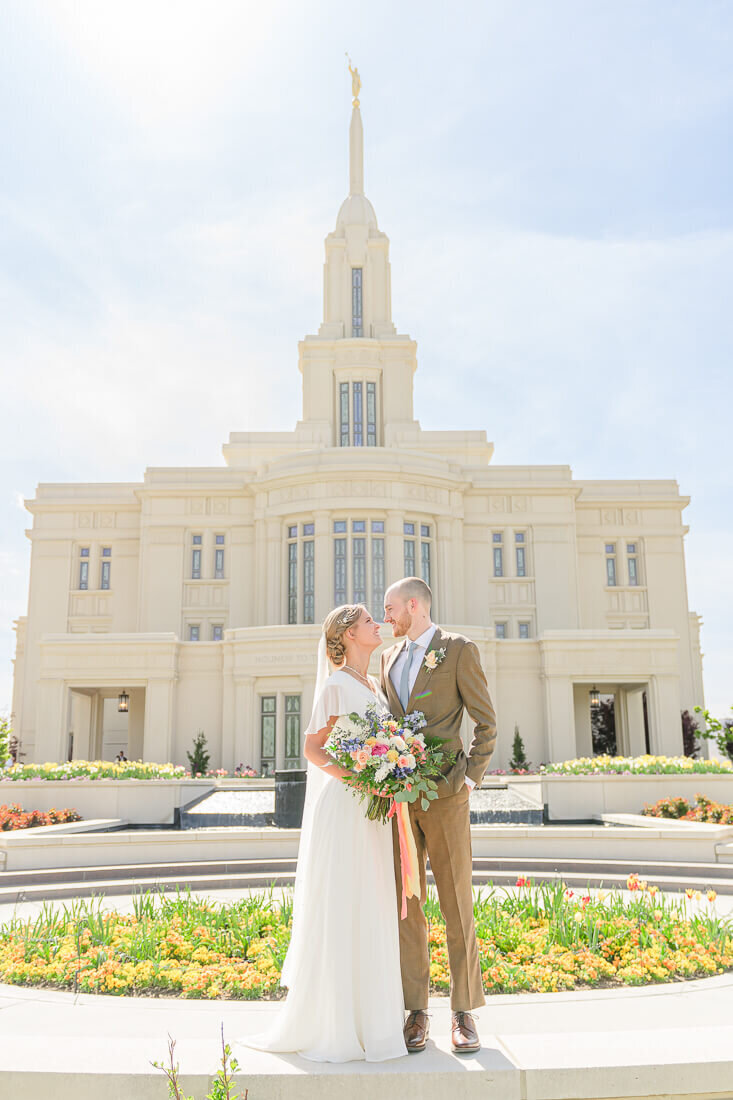 A summer bride holds a colorful bouquet with peach ribbons and looks up at her groom wearing a light brown suit with a pale blue tie with a full view of the Payson temple behind them