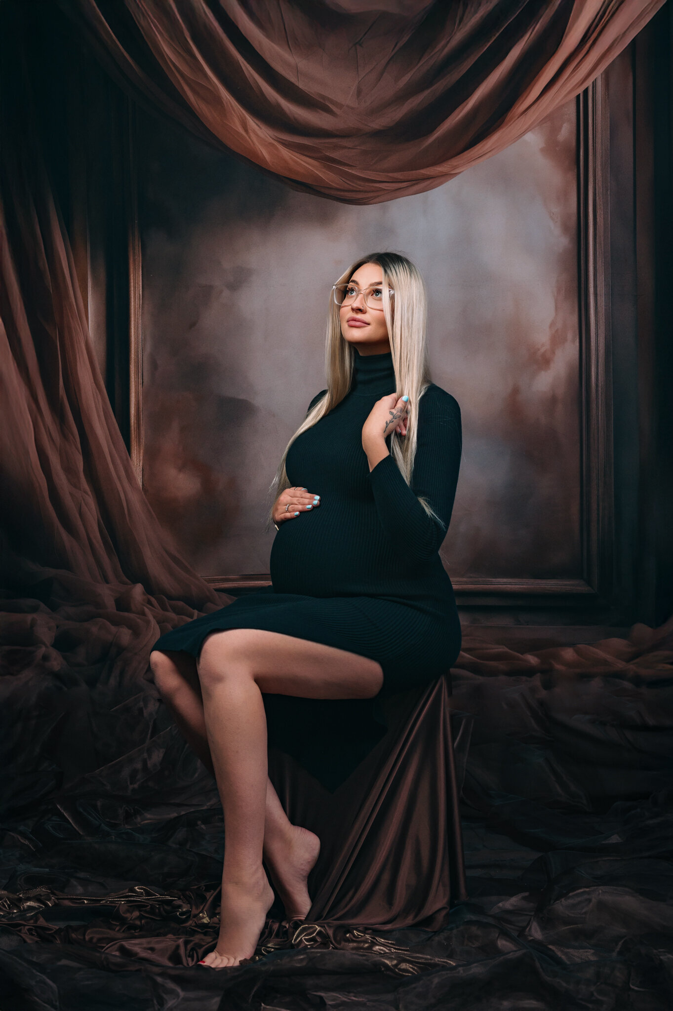 stunning blonde expecting mother seated in black dress in front of a brown tones fabric and large frame