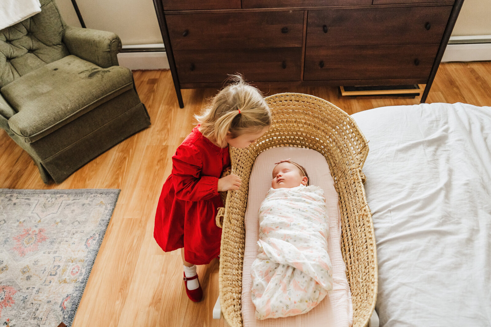 toddler girl peeks on tiptoe into bassinet at sleeping newborn sister