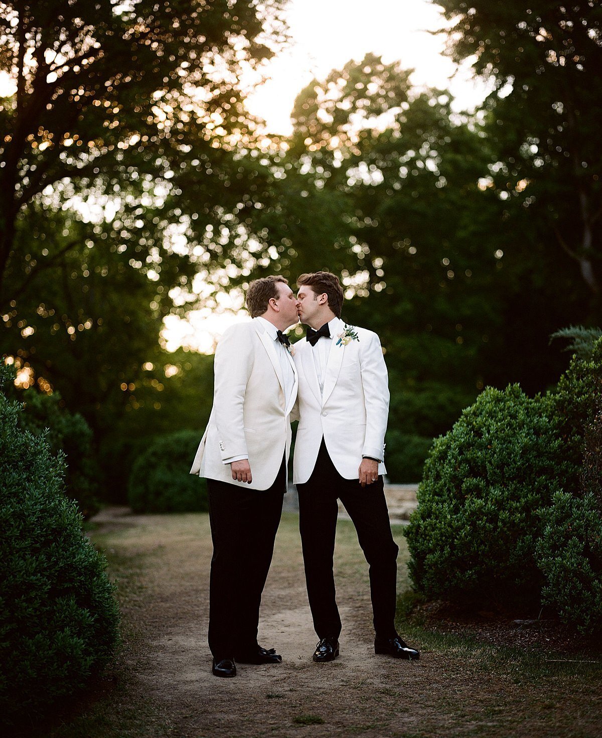 Two grooms wearing black pants and white tuxedo jackets kissing