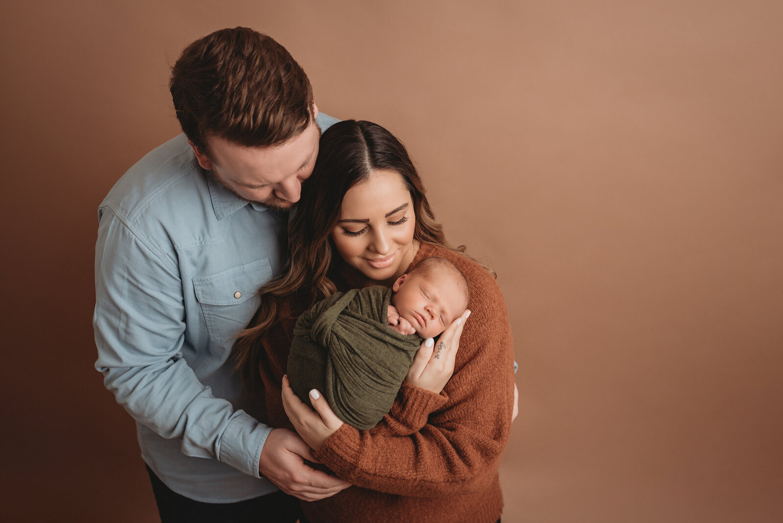 Newborn family portrait with mom holding baby. on tan backdrop. Family is wearing neutral colors of blue, brown and green.