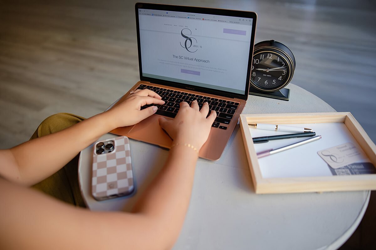 Overhead branding photo of a woman typing on a rose gold laptop at a minimalist desk setup with a modern clock, iPhone, and pens. Captured by a Seattle brand photographer, this image highlights a clean, professional workspace during a branding photoshoot for women entrepreneurs growing their personal brands.