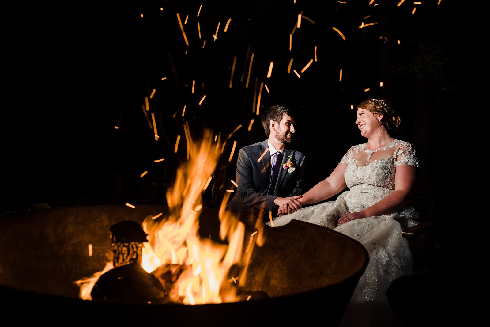 bride and groom sit at a bonfire outside of their wedding reception as sparks fly in the night sky