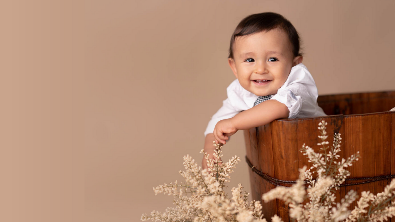 Smiling baby in a wooden tub, wearing a white shirt, surrounded by delicate beige flowers, set against a soft brown background. A joyful and warm scene.