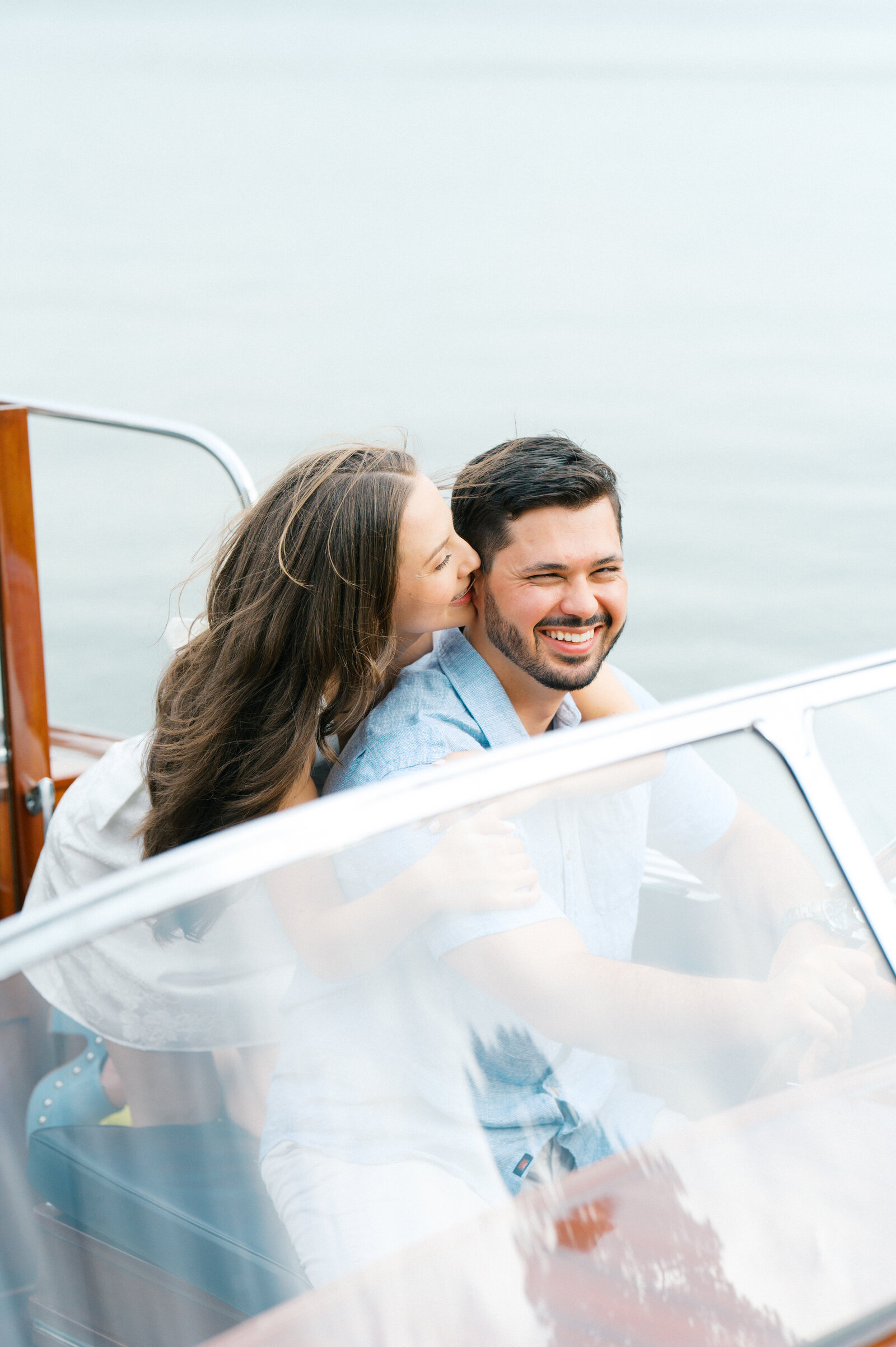Brunette girl smiles kissing her fiances cheek while he drives classic wooden boat on Lake Oconee