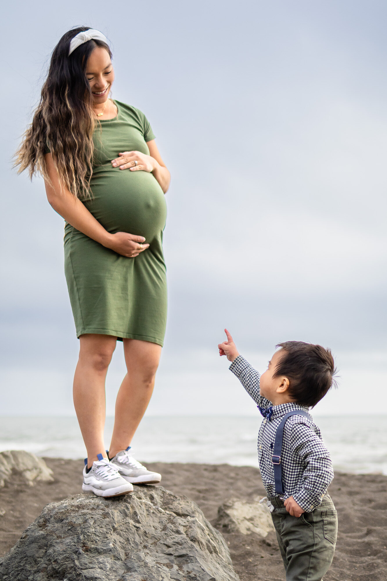 Maternity portrait of a mom smiling at her toddler son who is pointing up at her on a beach rock Maternity - Ellobelle Photography