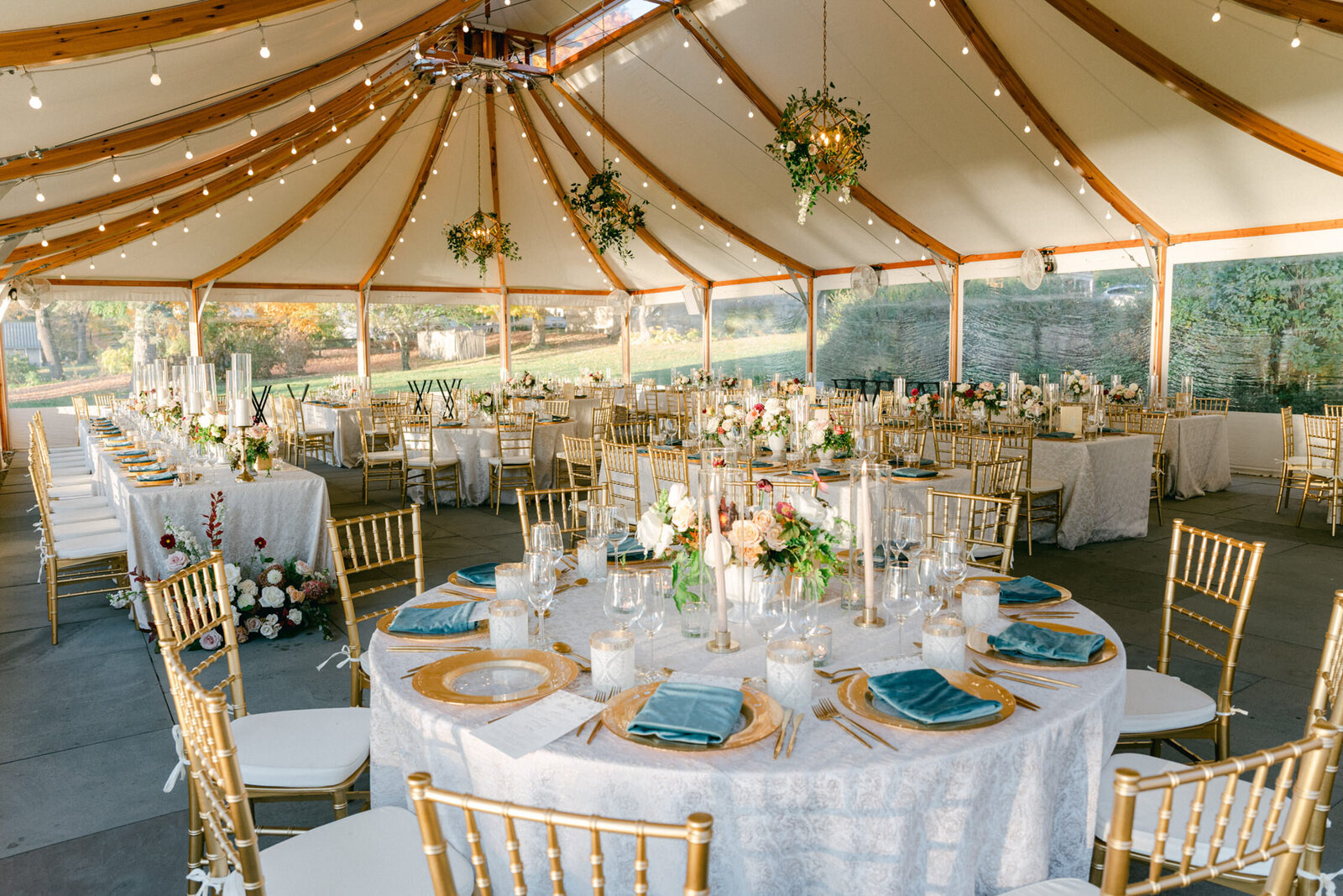 Tables are set under a sailcloth tent. Gold chairs are places around table with gold accessories and blue napkins on china.
