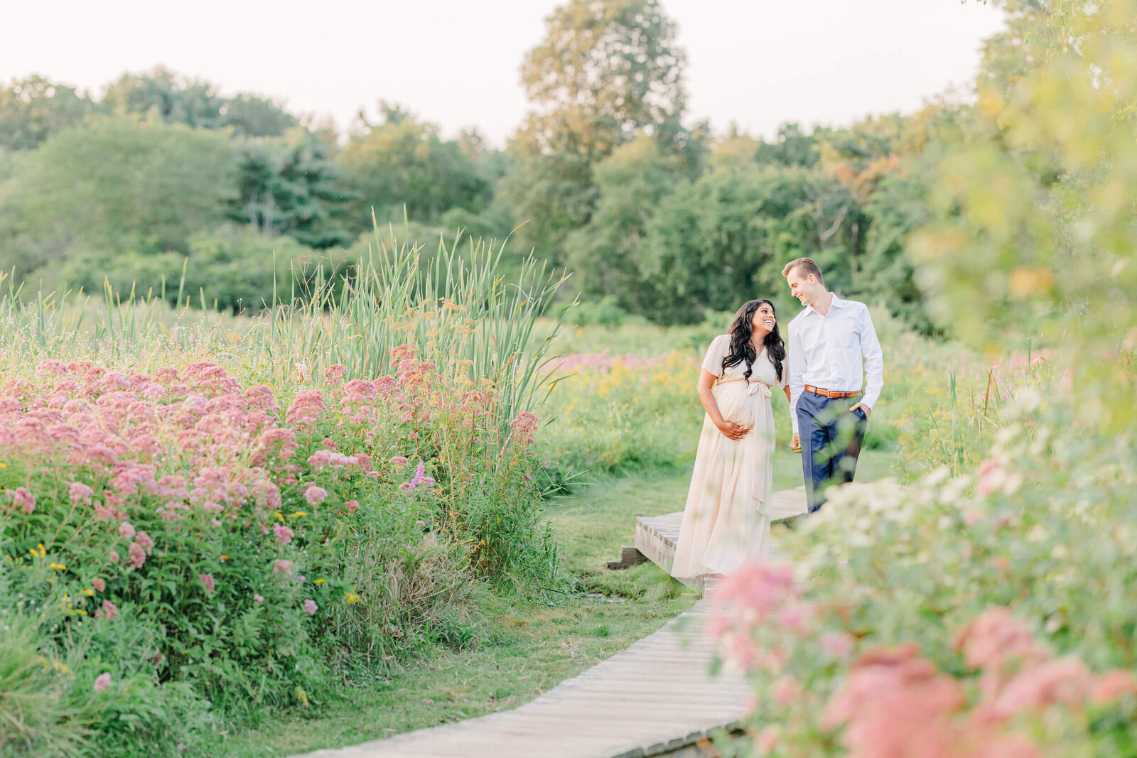 Pregnant woman and her husband walk together on a wooden path in a field of wild flowers