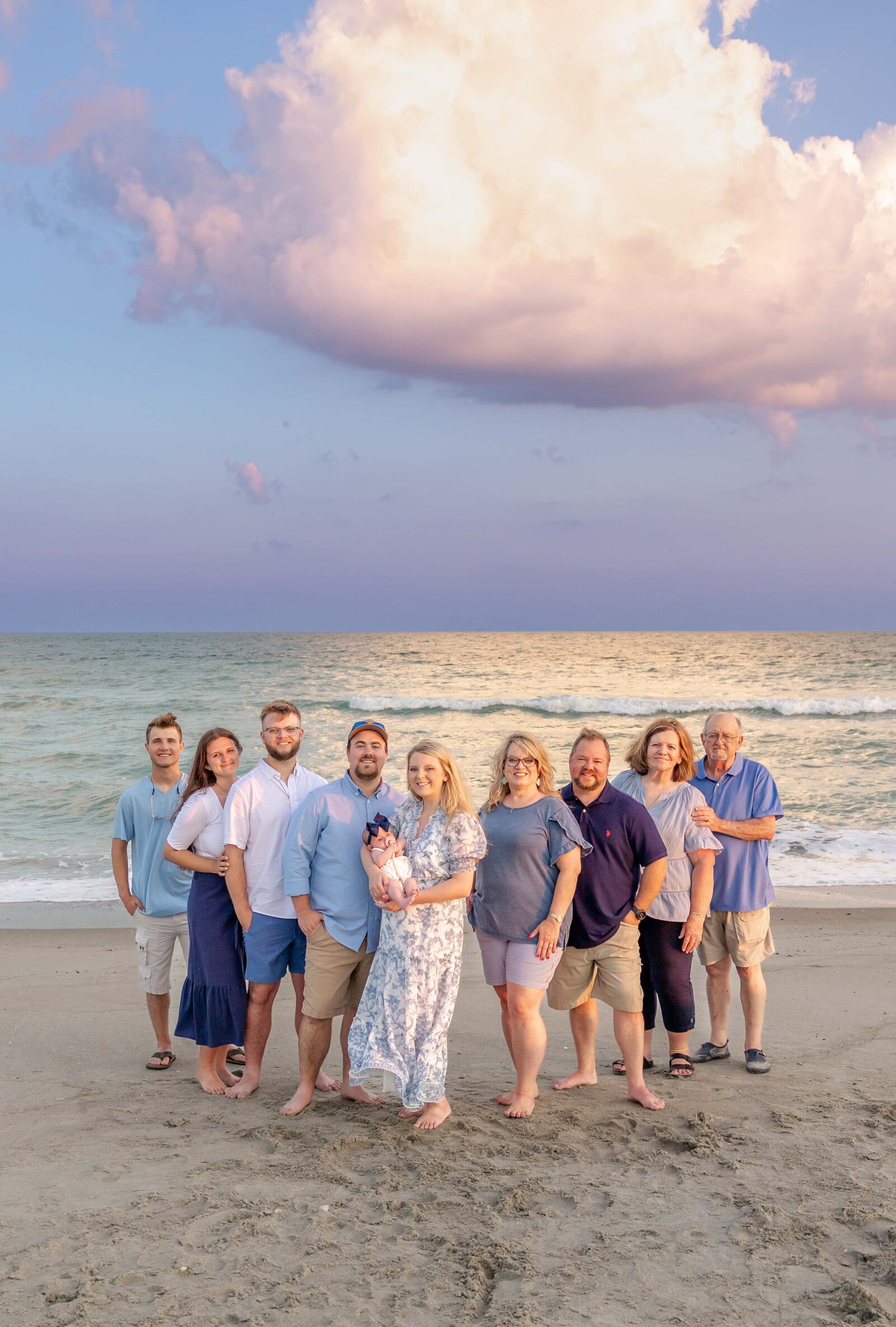 A family poses on the beach under a striking cloud formation at sunset. Dressed in blue and pastel outfits, they capture a warm, relaxed moment by the ocean. Perfect for North Carolina family photography, highlighting stunning beachside gatherings.