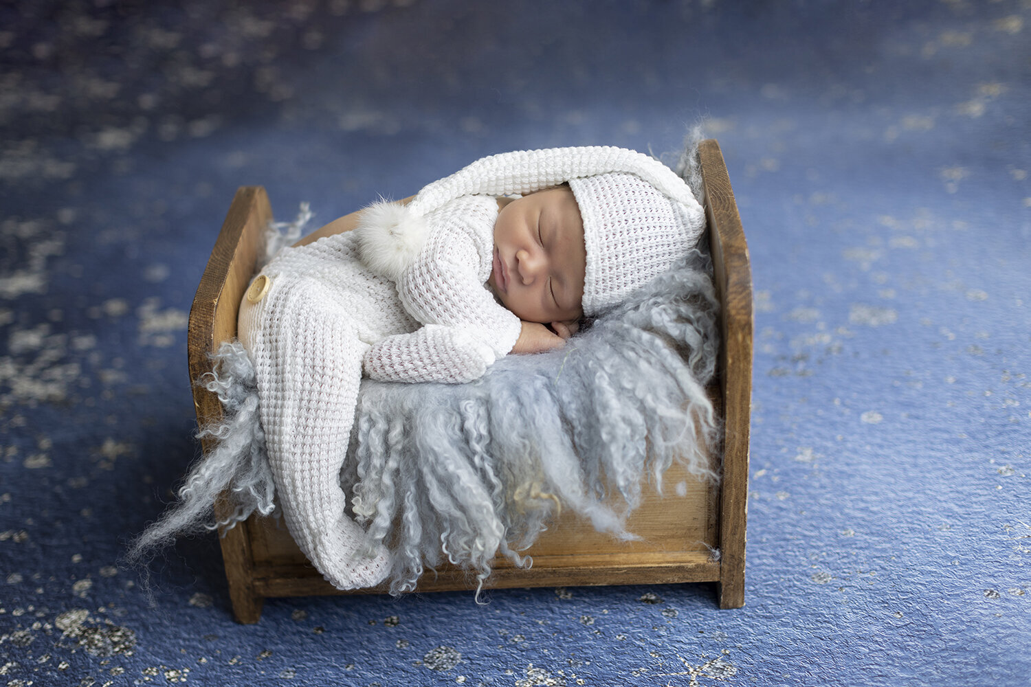 Newborn boy wears white sleep suit poses on tiny bed