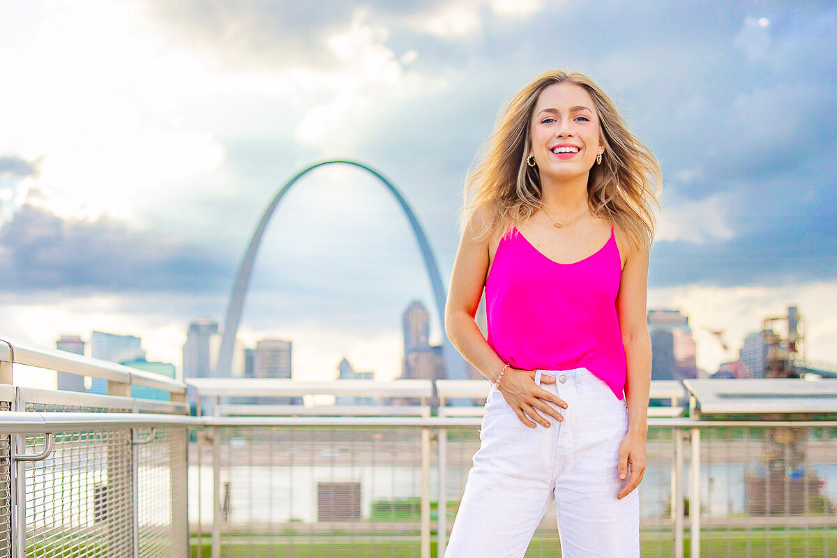 senior smiling in a wide stance with the arch and st. louis skyline behind her