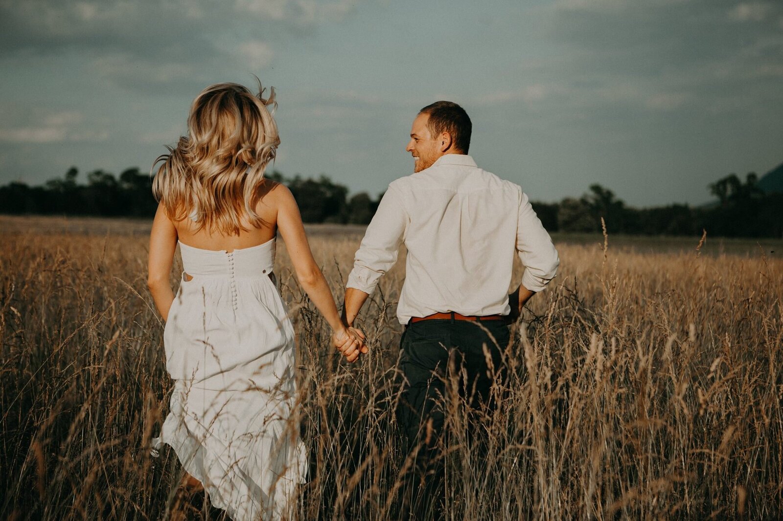 A man and a blond woman run through a field of long grass at sunset