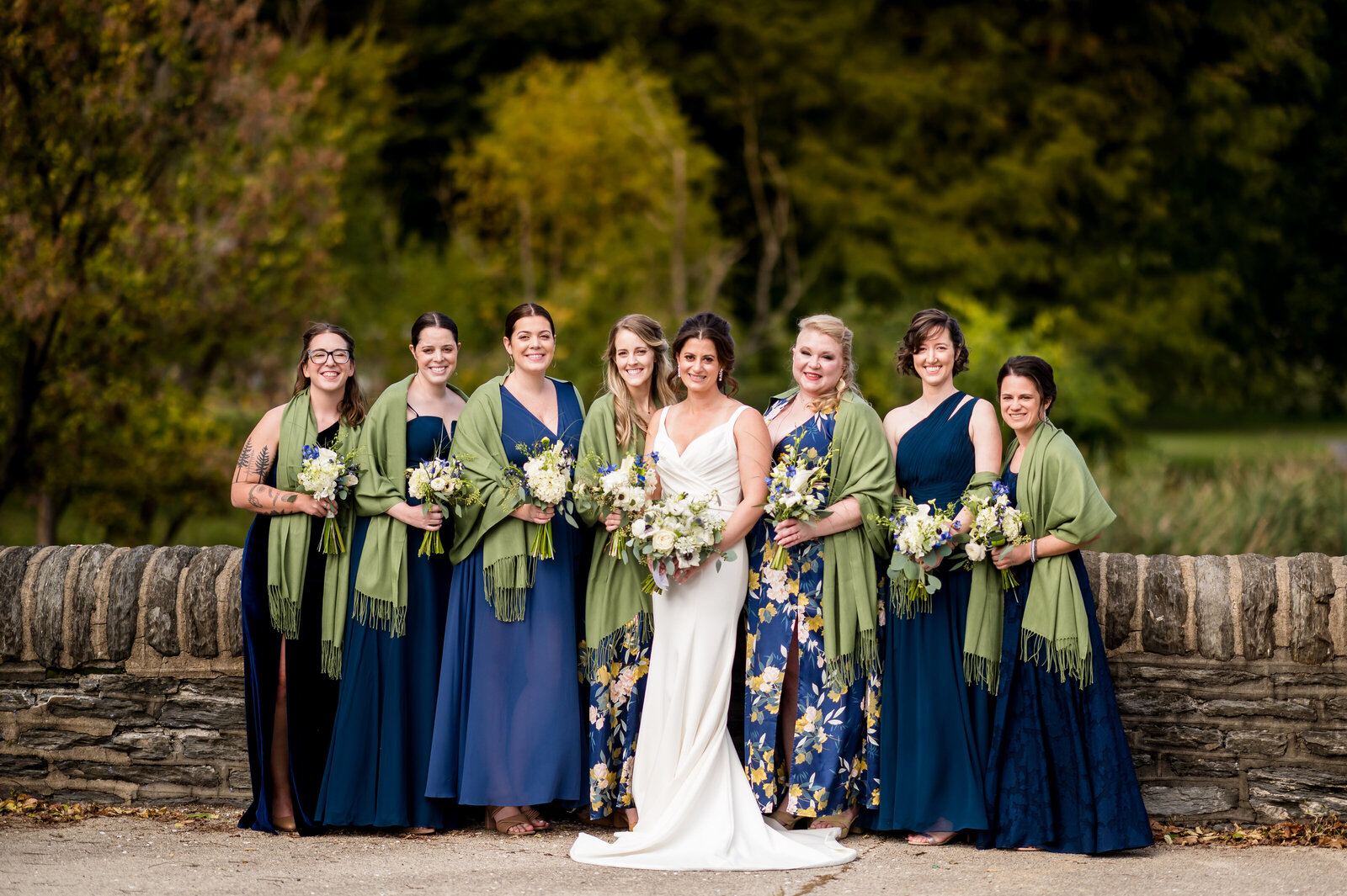 A bride in a white dress stands with seven bridesmaids wearing blue dresses and green shawls, holding bouquets, in front of a stone wall with a lush, green background.