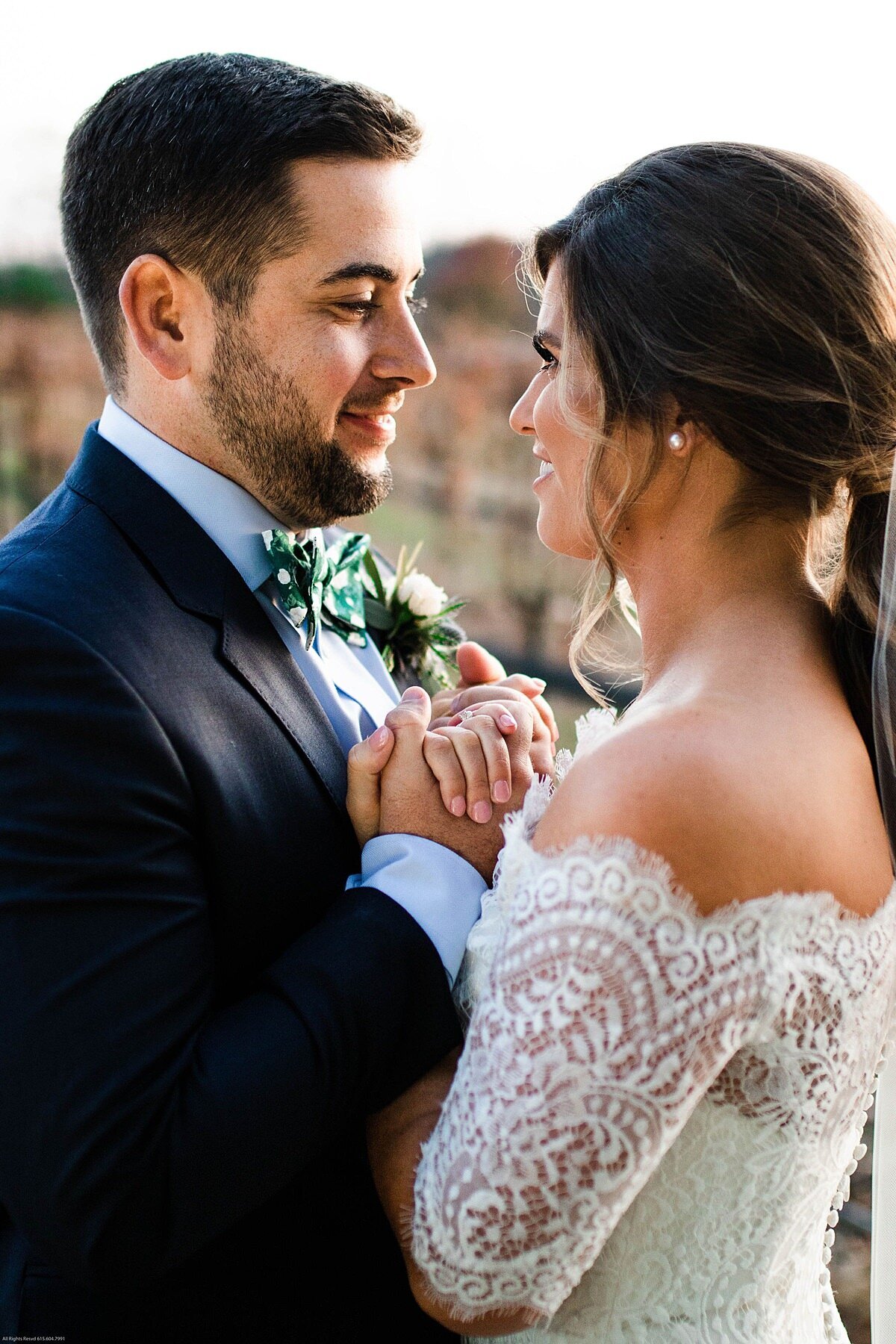 bride in a long sleeved white lace dress holds hands with the groom wearing a dark blue suit as they look into each other's eyes