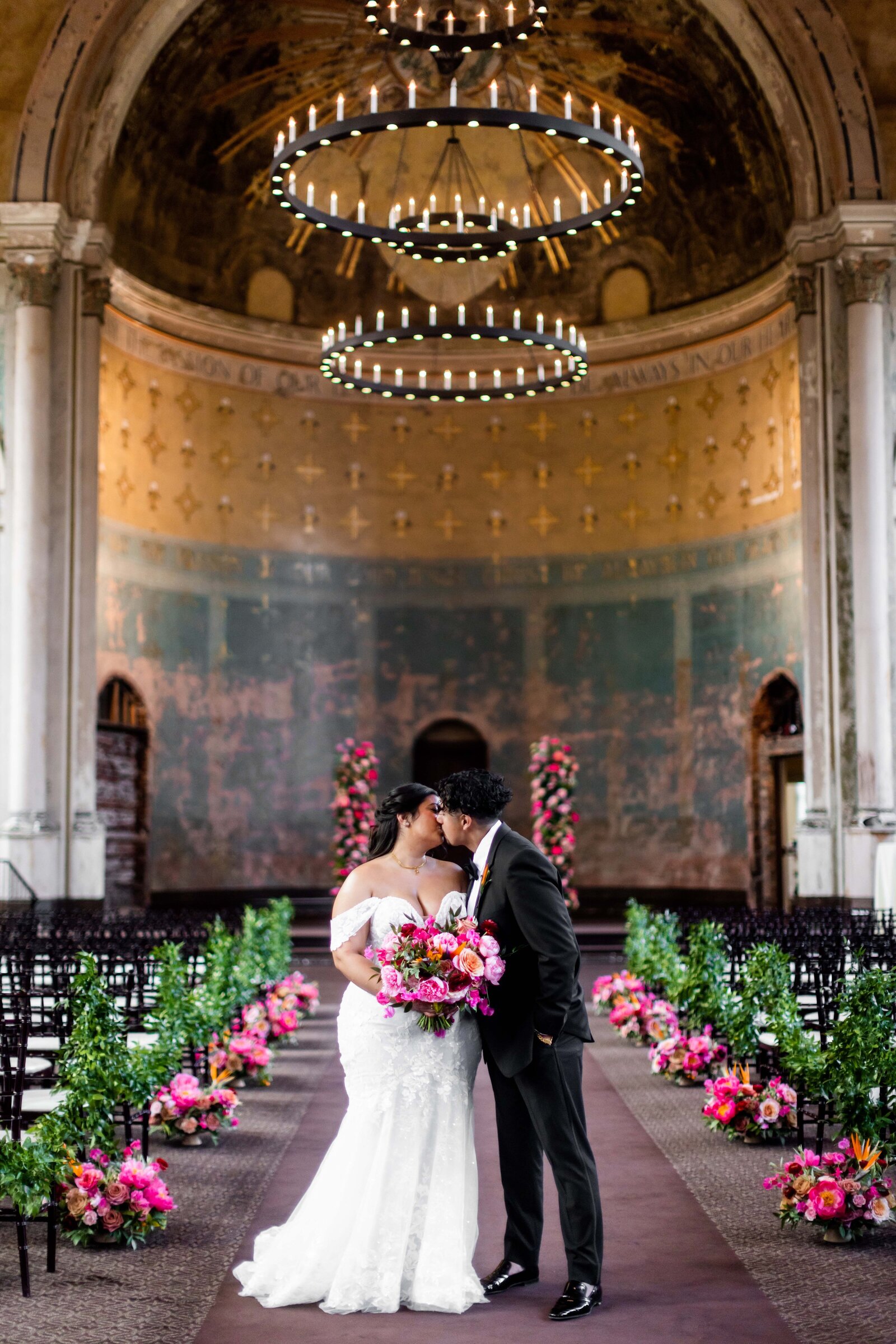 This enchanting photograph features Mary Grace and Martin standing hand-in-hand, enveloped in a dreamy aisle of vibrant pink florals during their wedding ceremony. The aisle is lavishly decorated with various shades of pink flowers, creating a romantic and lush pathway that leads to the couple. The stunning visual composition captures the essence of their love and the joy of the moment, enhanced by the floral splendor that surrounds them. This image is a testament to the couple's vibrant personality and their celebration's exquisite attention to detail, making it a captivating highlight of their wedding day. Ideal for couples seeking inspiration for creating a visually impactful floral arrangement on their special day.