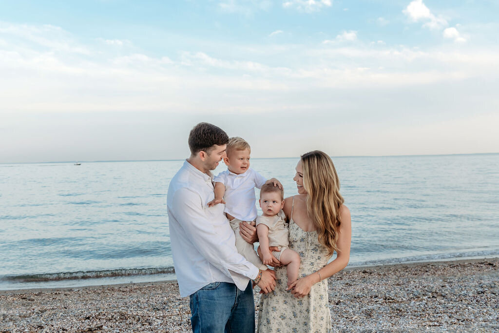 Connecticut Family Photographer captures a tender beach moment with parents holding their two young sons against a serene waterfront backdrop during golden hour