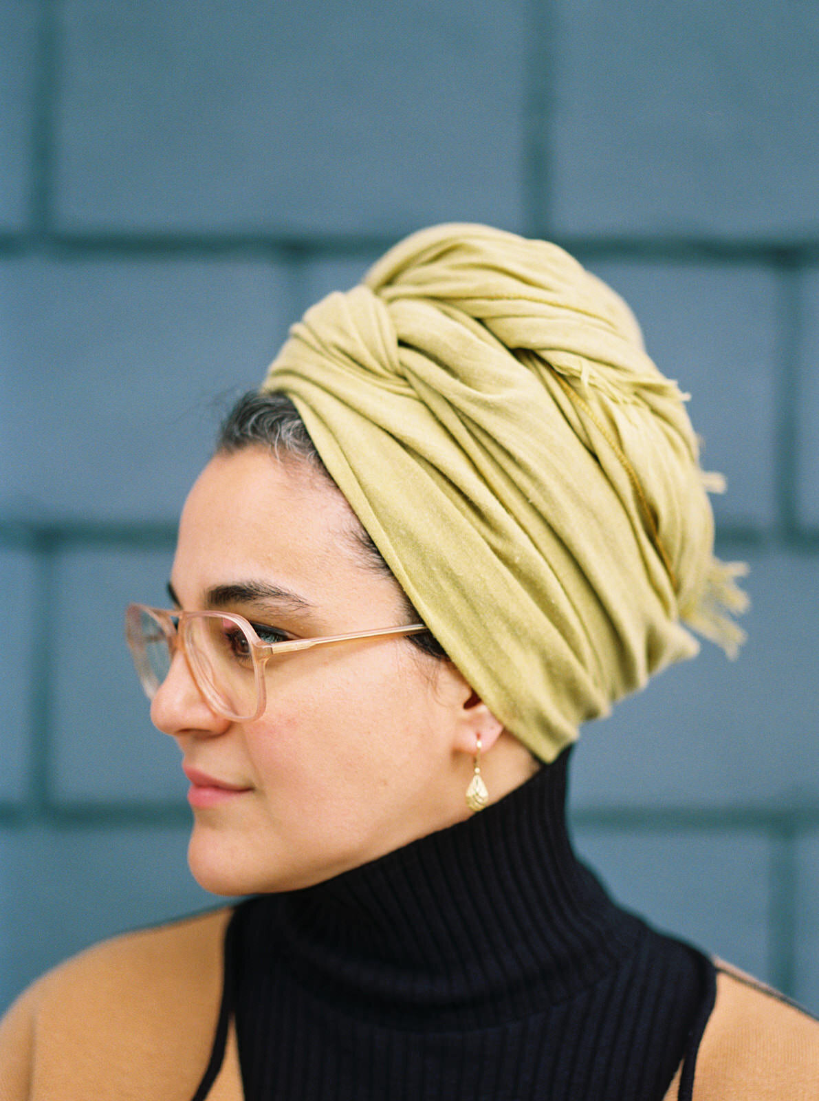 Side profile of a woman wearing a green headwrap and glasses, standing against a bold teal wall.