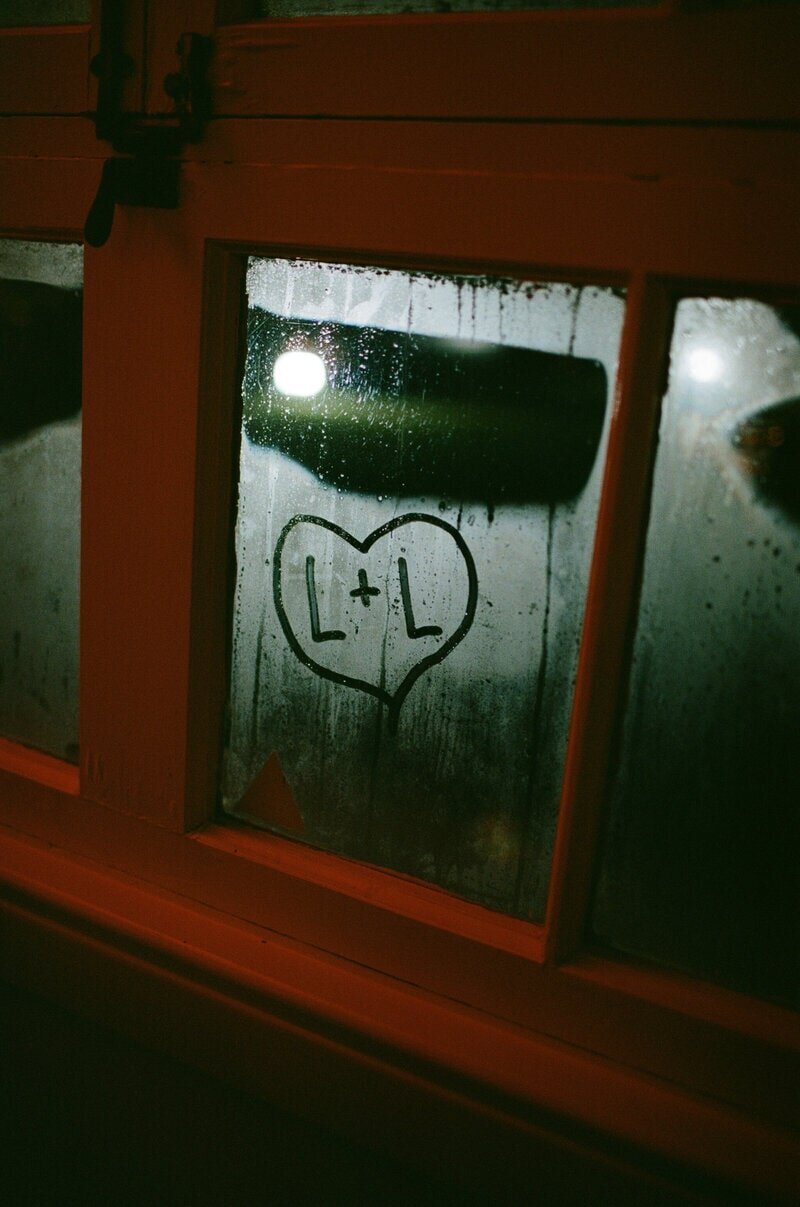 A foggy window with a heart drawn on it, containing the letters L + L. The background is dimly lit, with a light source partially visible through the condensation on the glass.