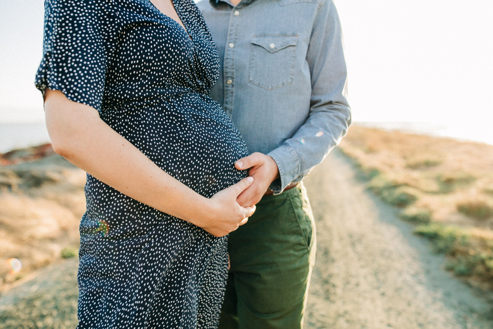 closeup of a pregnant belly in a navy dress with white dots while husband stands behind and has a hand on her belly