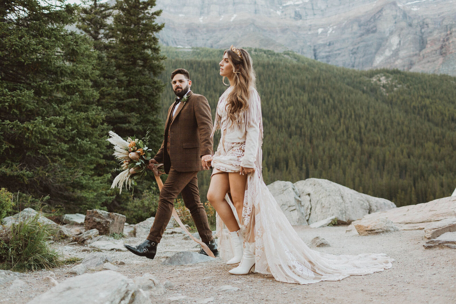 groom holding bouquet with bride in long veil walking among Canadian Rockies